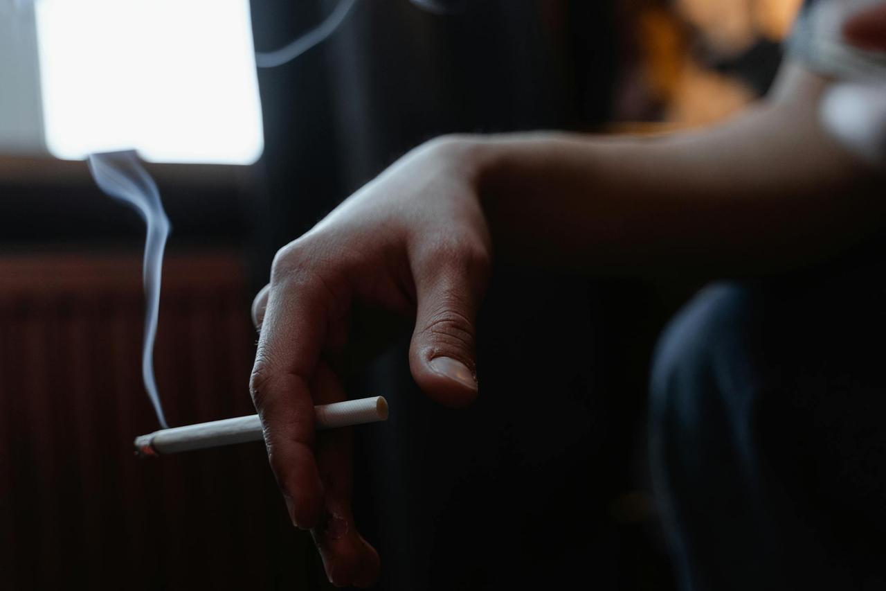Close-up of a hand holding a lit cigarette indoors with smoke rising in a dimly lit room.
