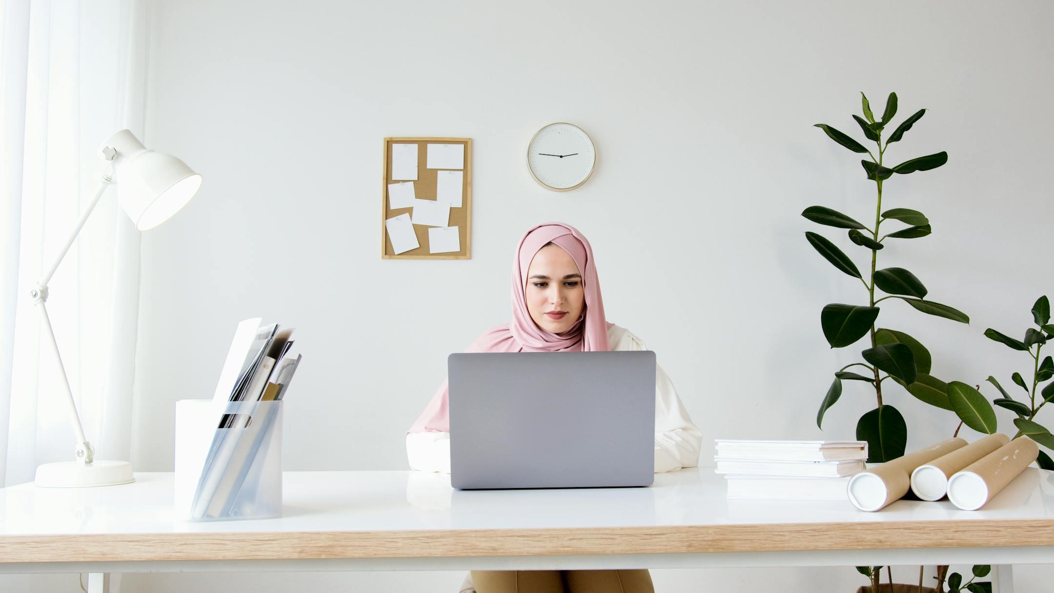 A Muslim woman in a hijab working on a laptop in a modern, minimalist office setting.