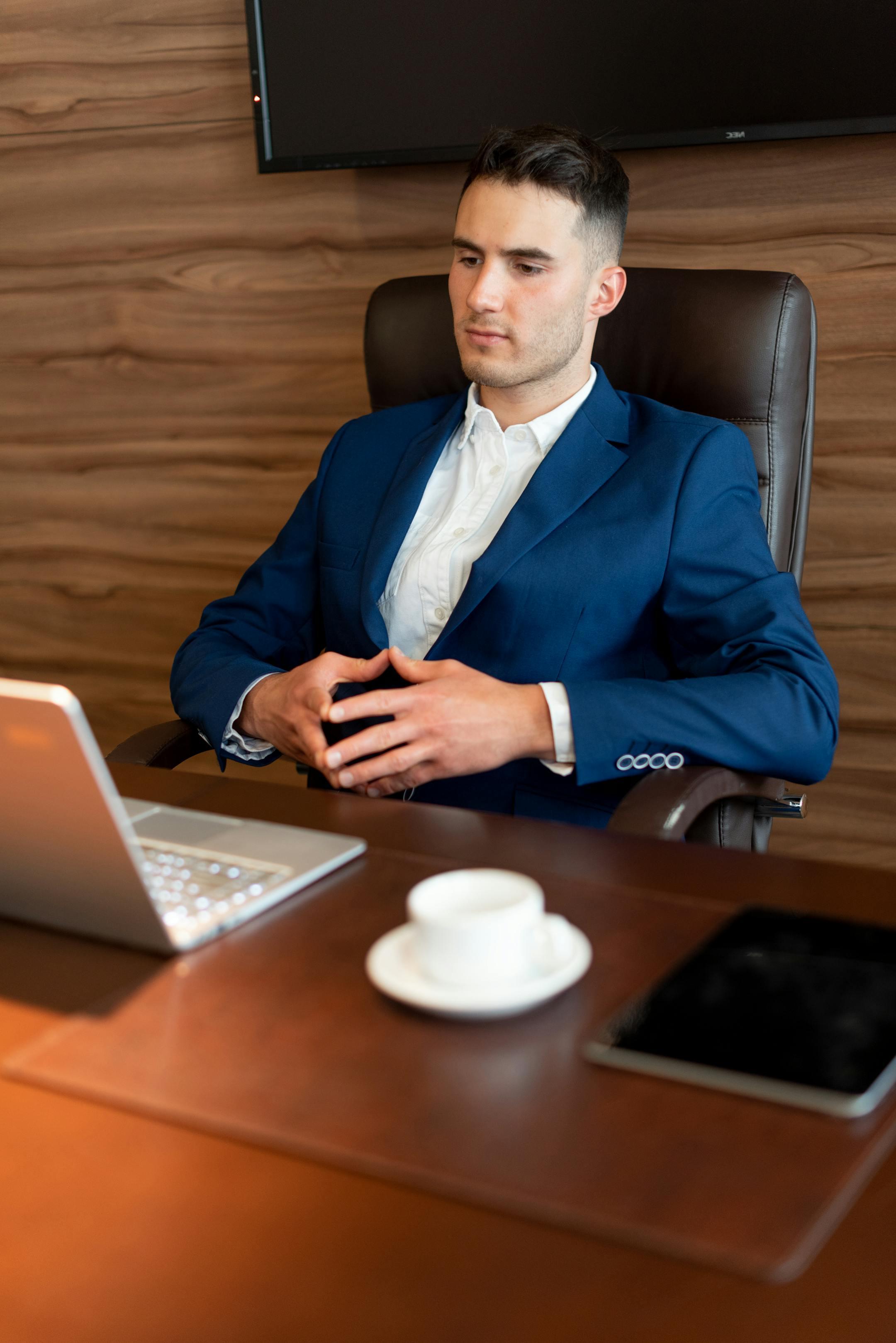 Professional businessman in a blue suit working at a desk with a laptop and coffee in a modern office setting.