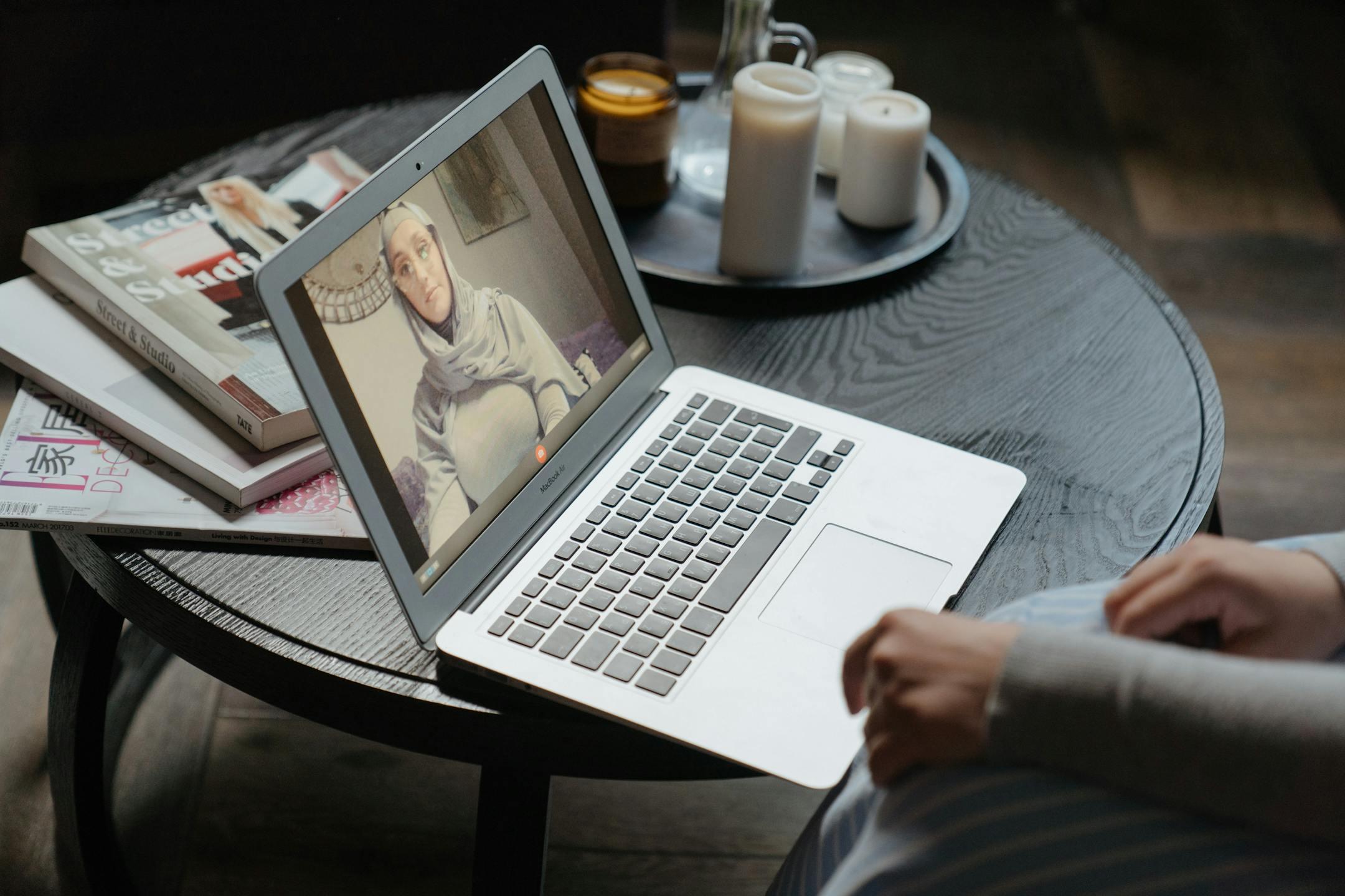 A woman in a hijab participating in a video call on a laptop, seated at a round table indoors.