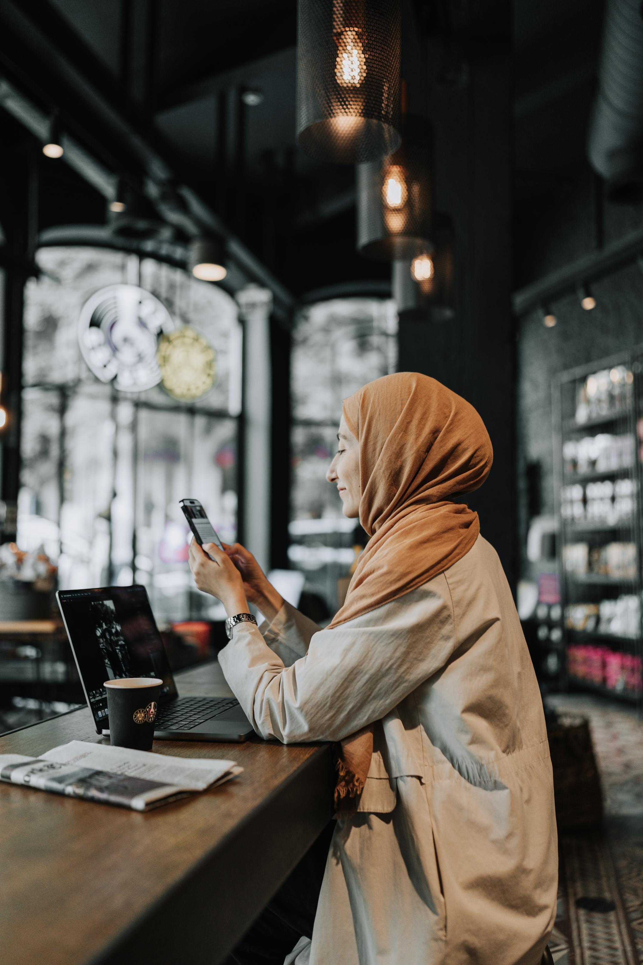 Female entrepreneur in a hijab multitasking in a cozy café. Working remotely on a laptop while checking her smartphone.