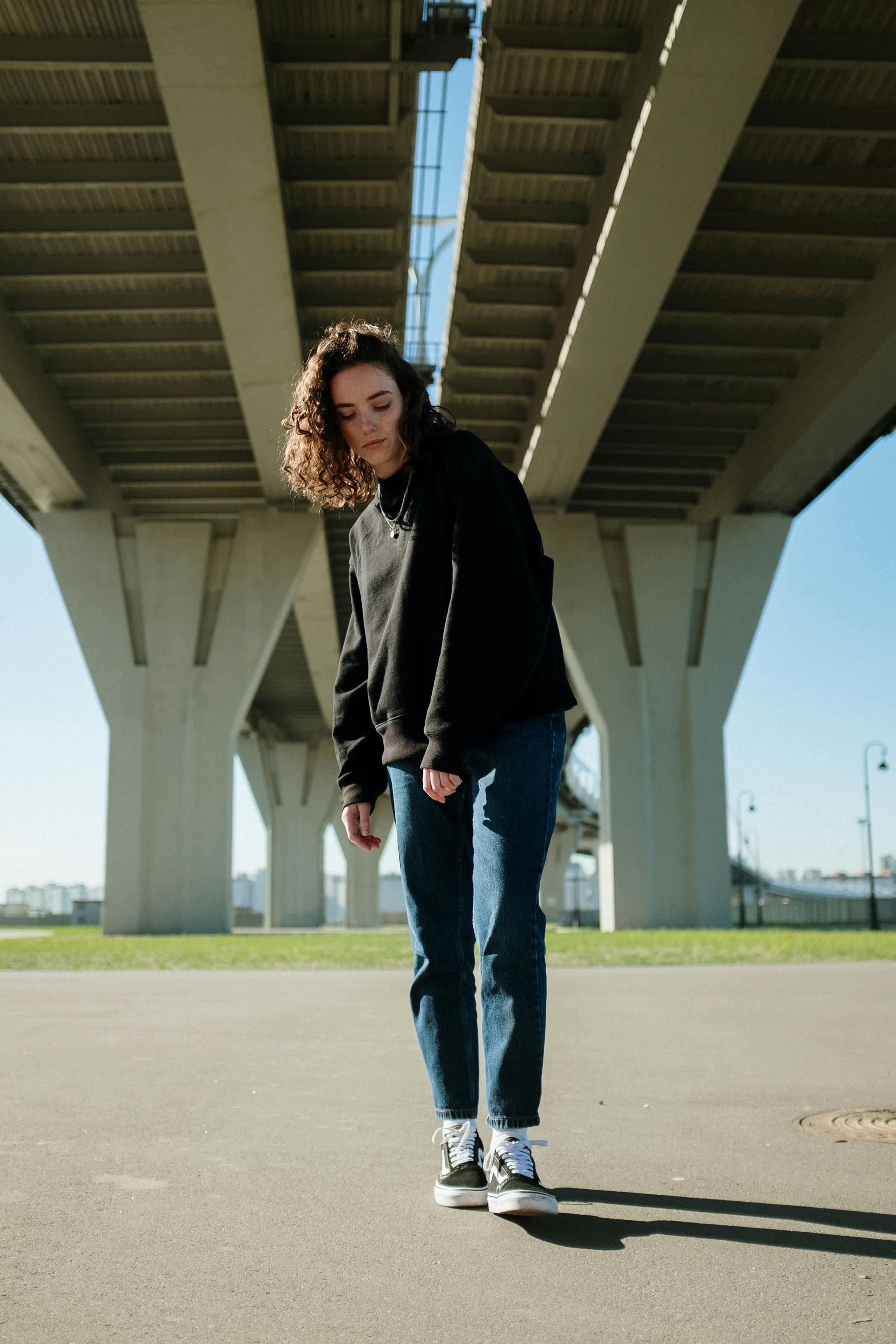 Young woman with curly hair stands under a modern urban bridge in daylight.