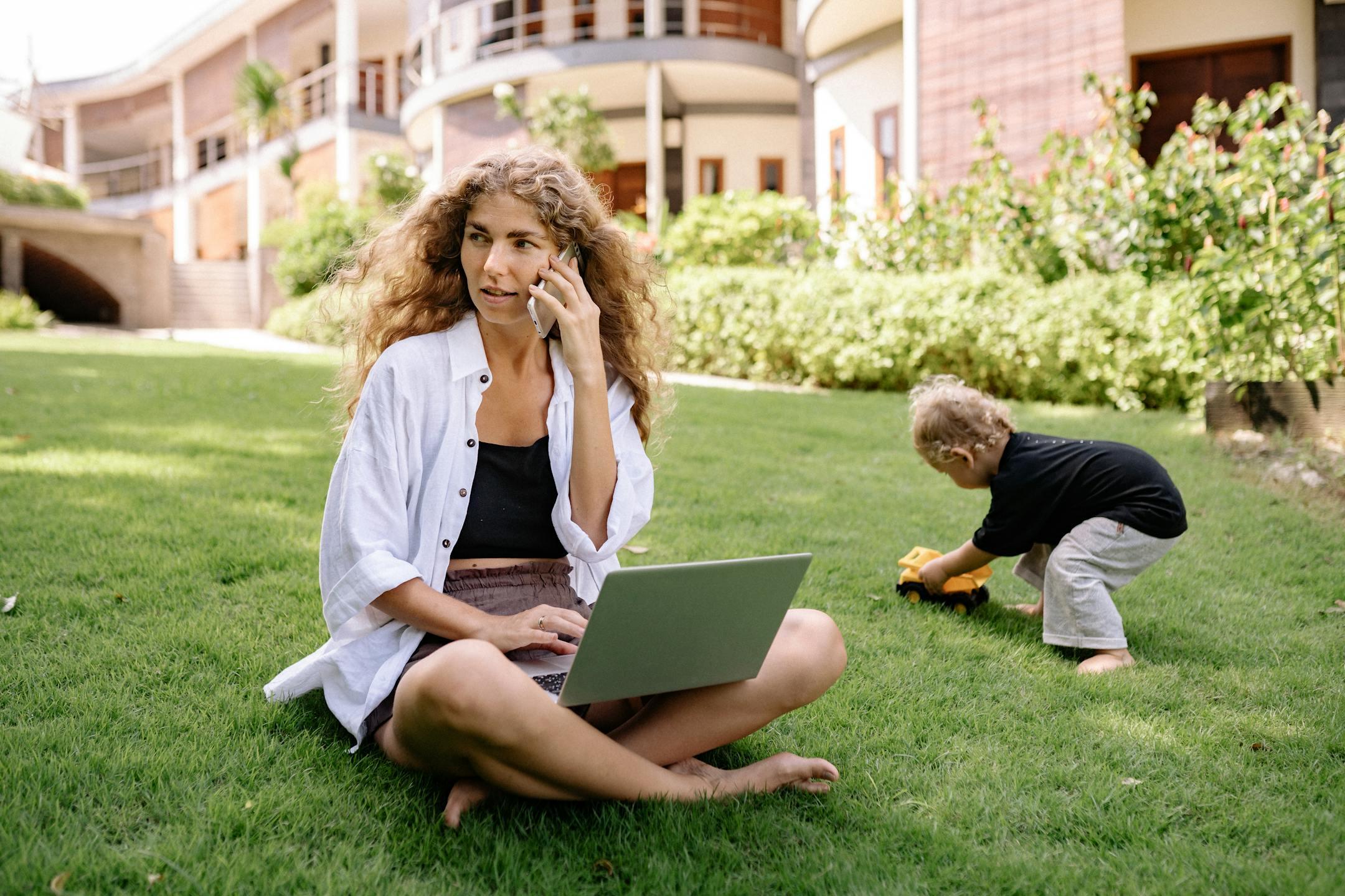 Woman multitasking on phone and laptop while child plays outdoors.