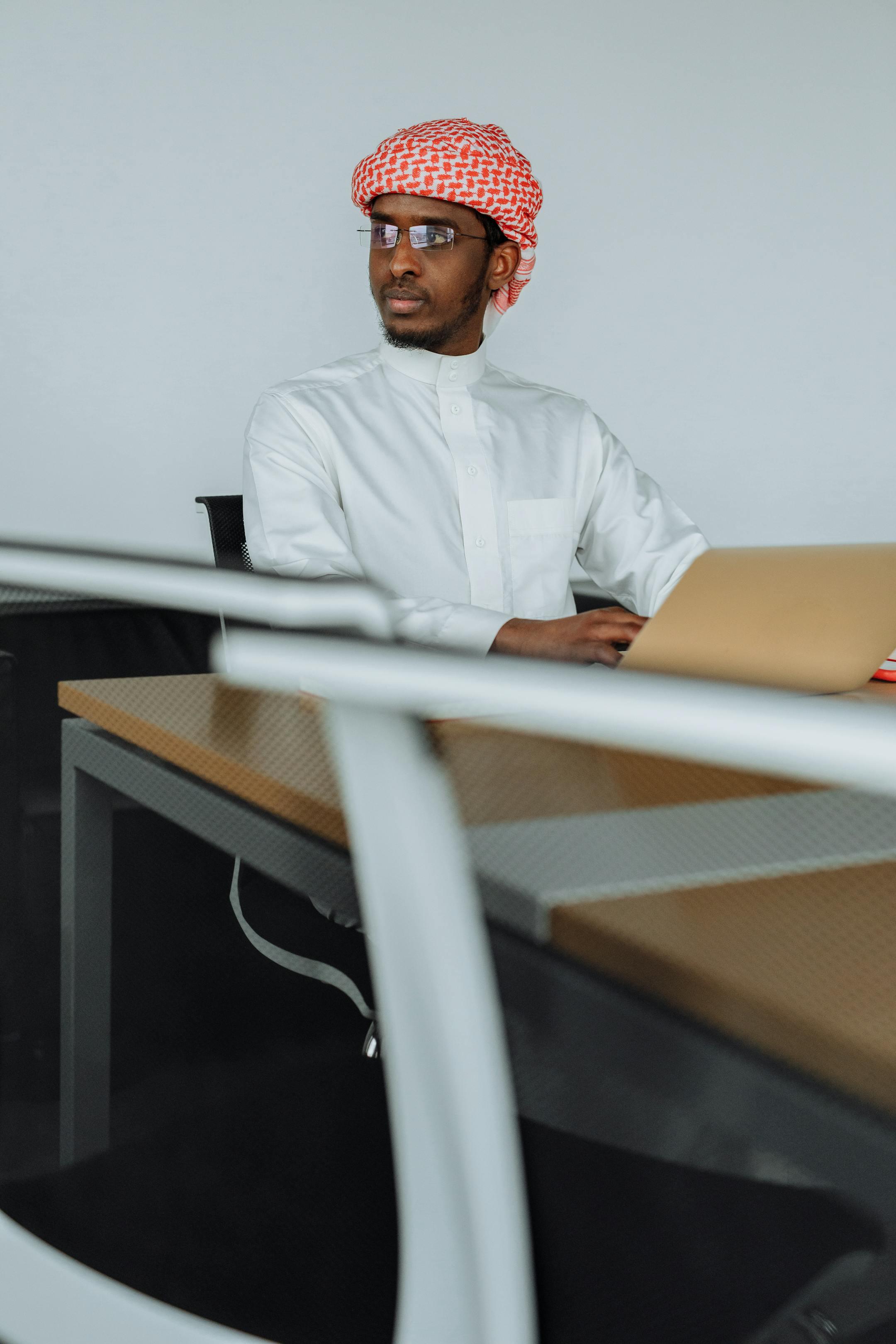 Middle Eastern man in traditional attire using laptop in modern office setting.