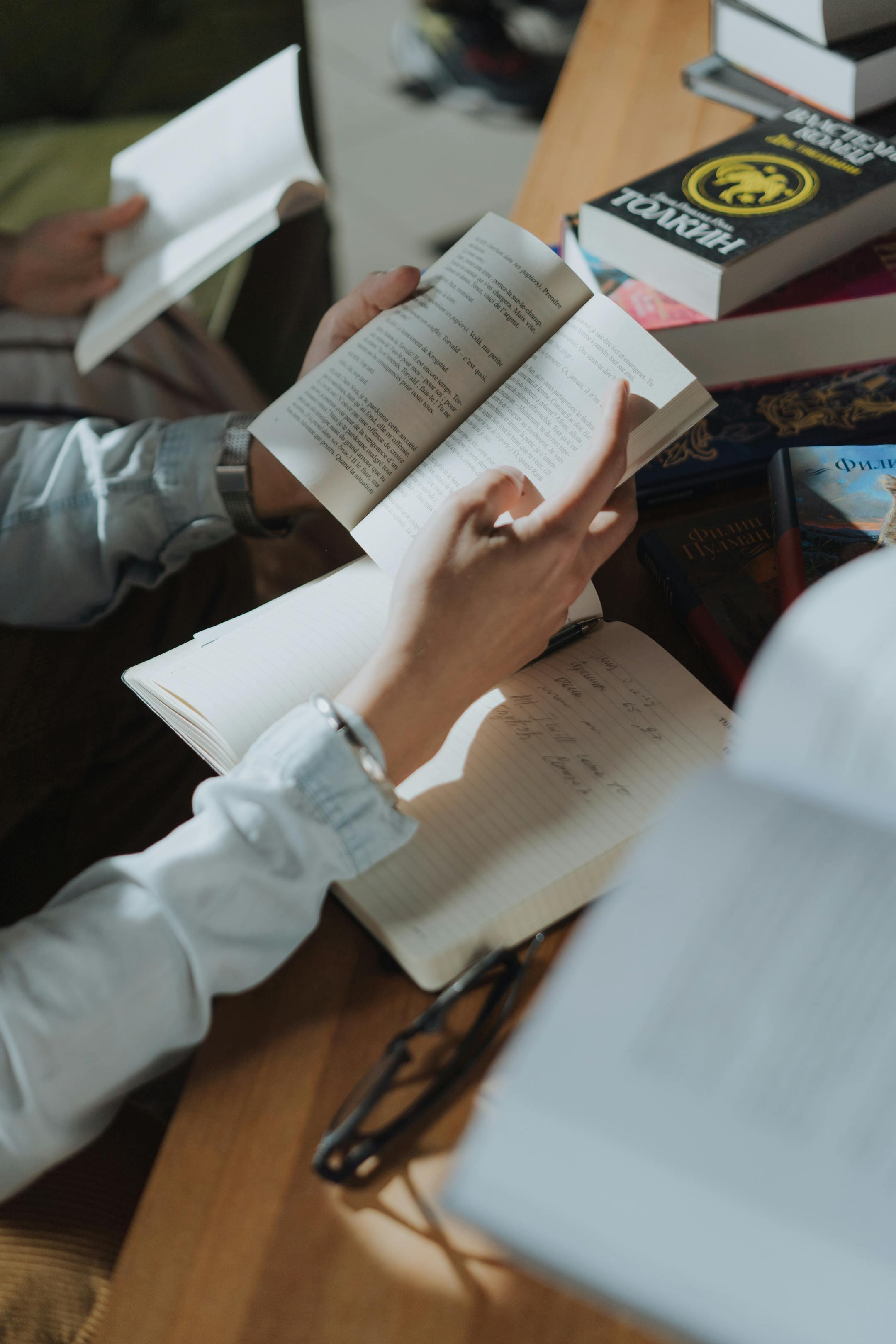 Person writing in a notebook while reading, surrounded by books in a cozy study setting.