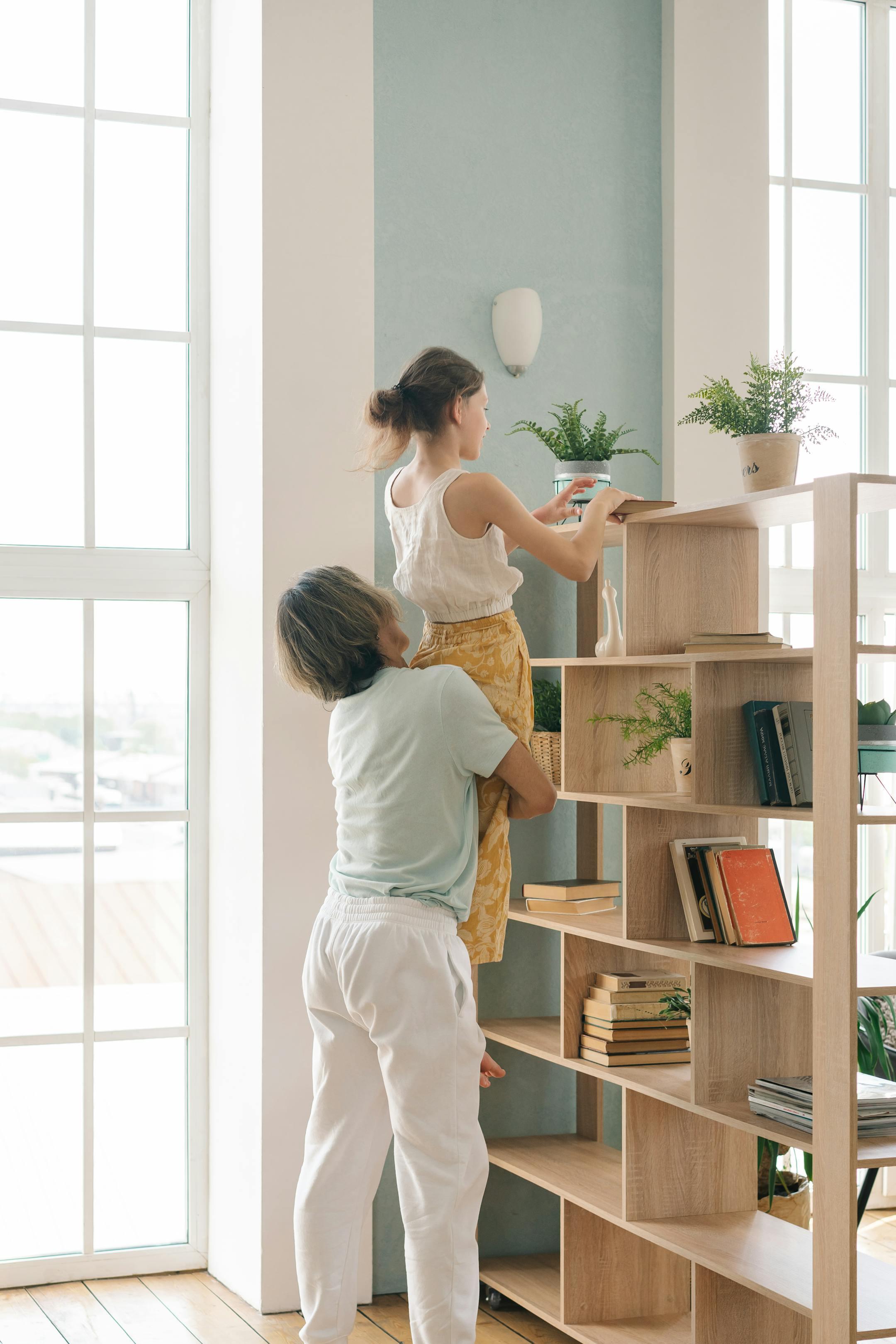 A father lifts his daughter to help her organize a bookshelf at home, fostering teamwork and family bonding.