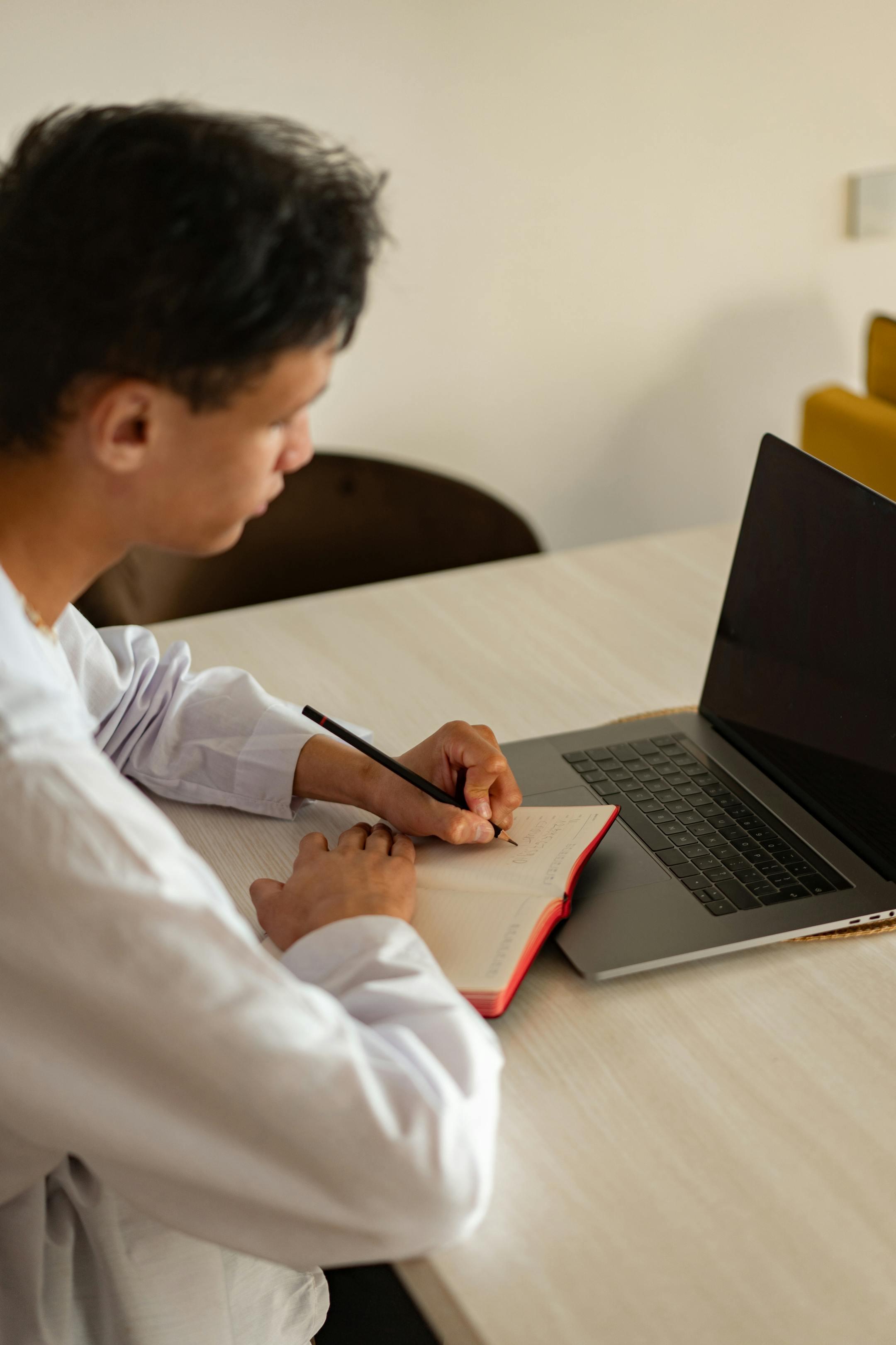 A young man focusing on his studies at home, using a laptop and notebook.