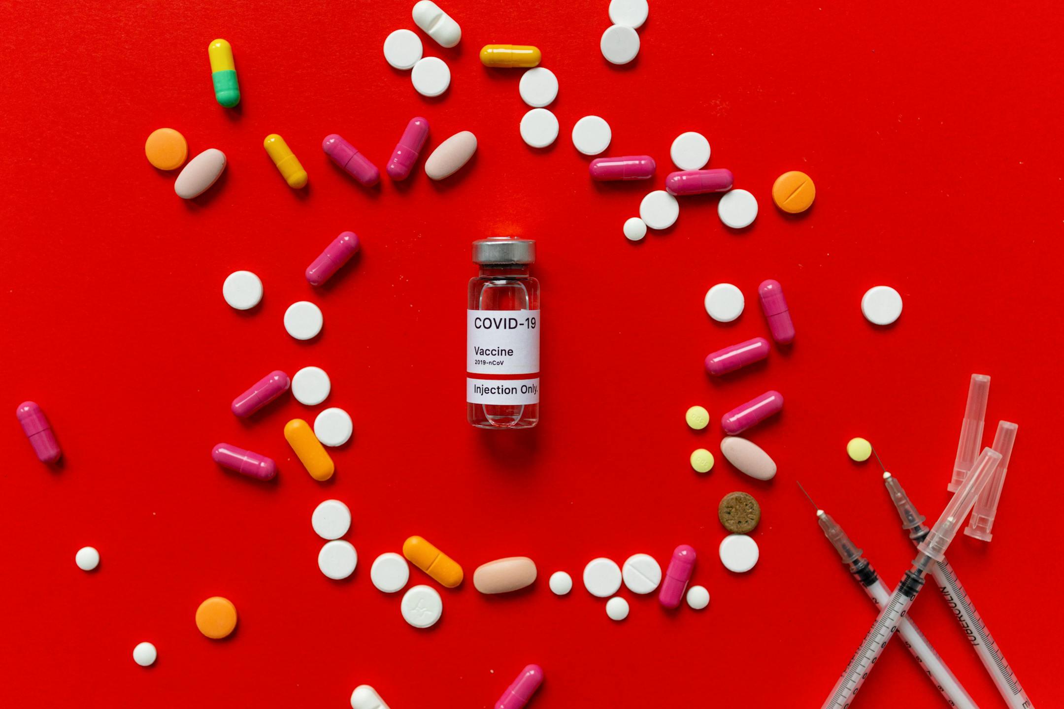 Colorful tablets and COVID-19 vaccine vial on a red background with syringes, symbolizing healthcare.