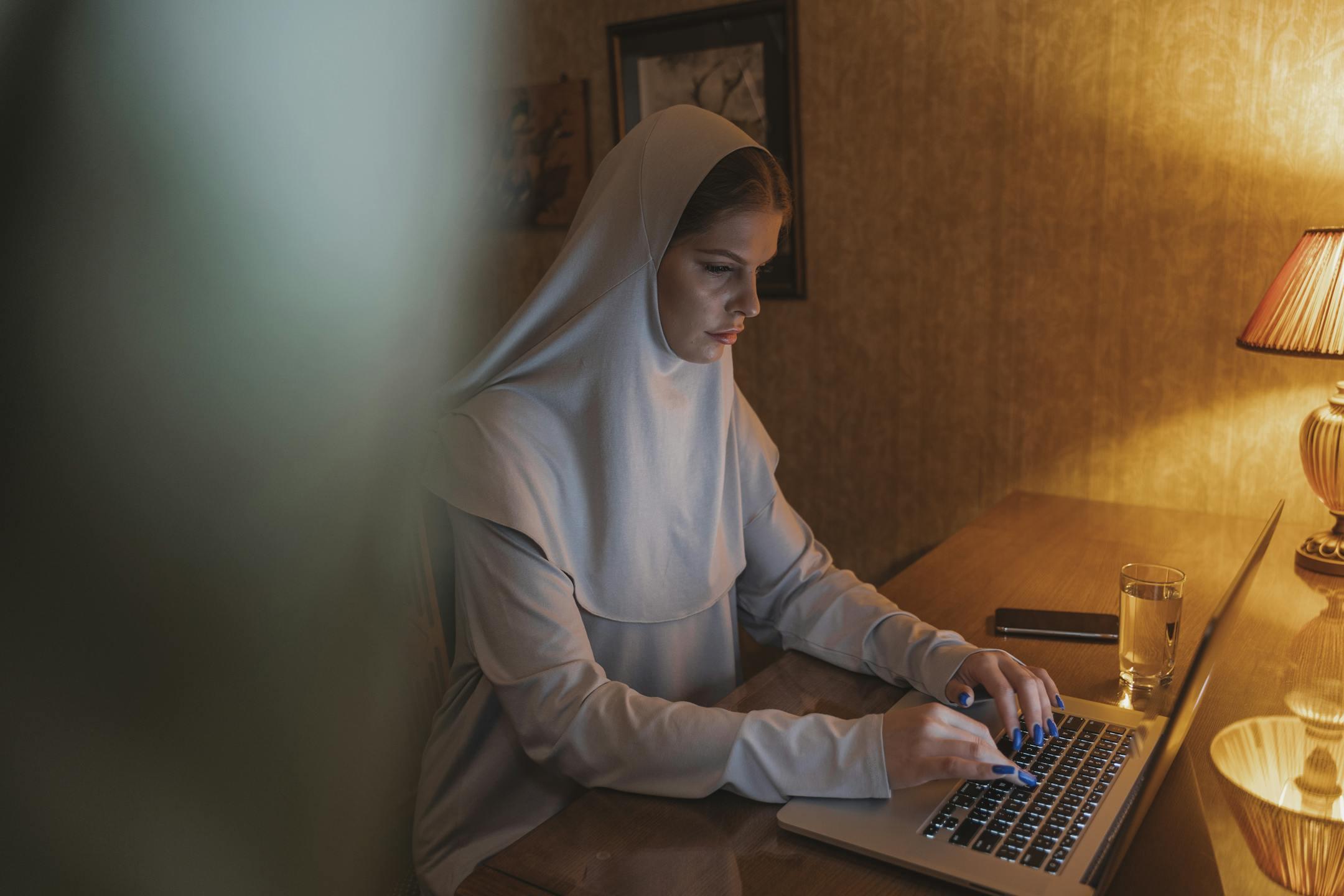 A woman in hijab focused on her laptop in a cozy home office setting.