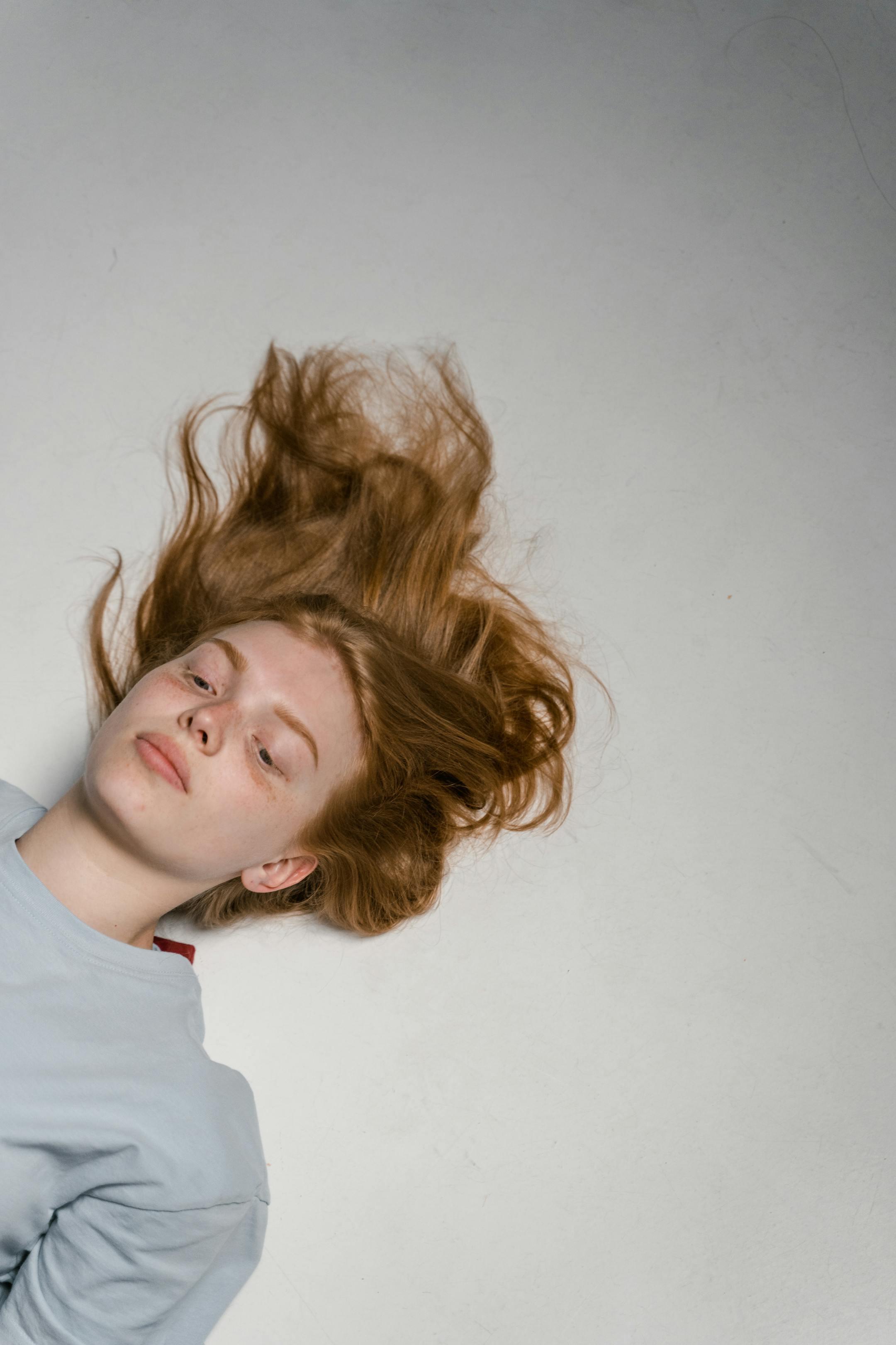 A woman with red hair lies on a light surface, conveying solitude and introspection.