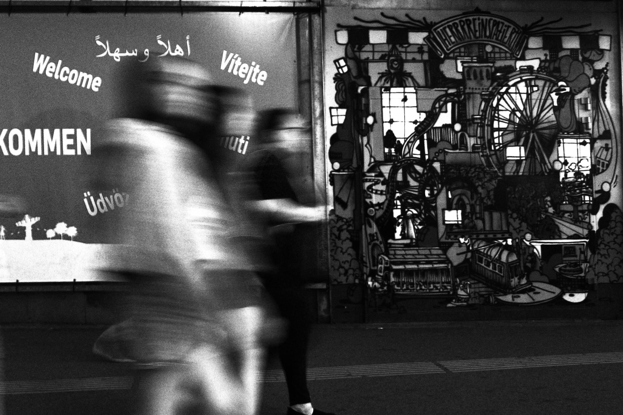 Blurred people walking past a multilingual welcome sign and mural in an urban setting.
