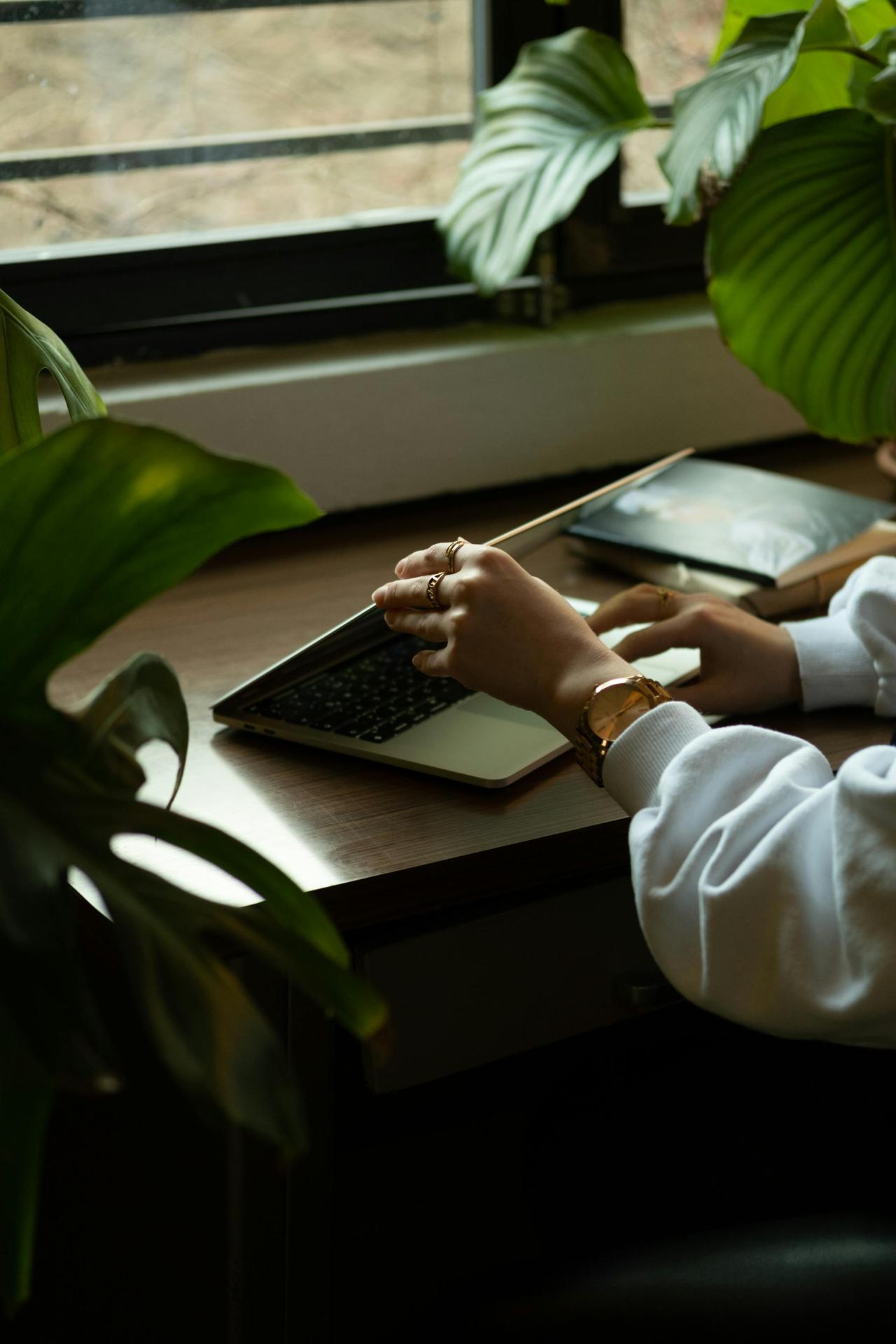 A woman working remotely on a laptop by a window, surrounded by lush houseplants.