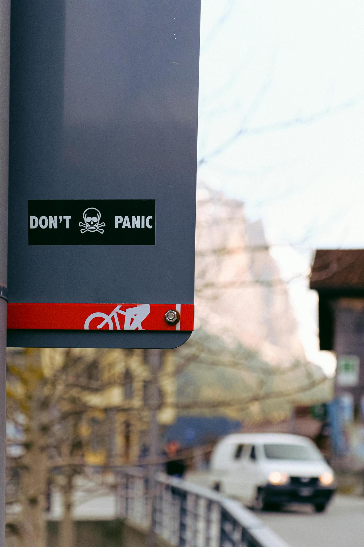 Close-up of sign with 'Don't Panic' message in urban setting, featuring vehicle and bike symbol.