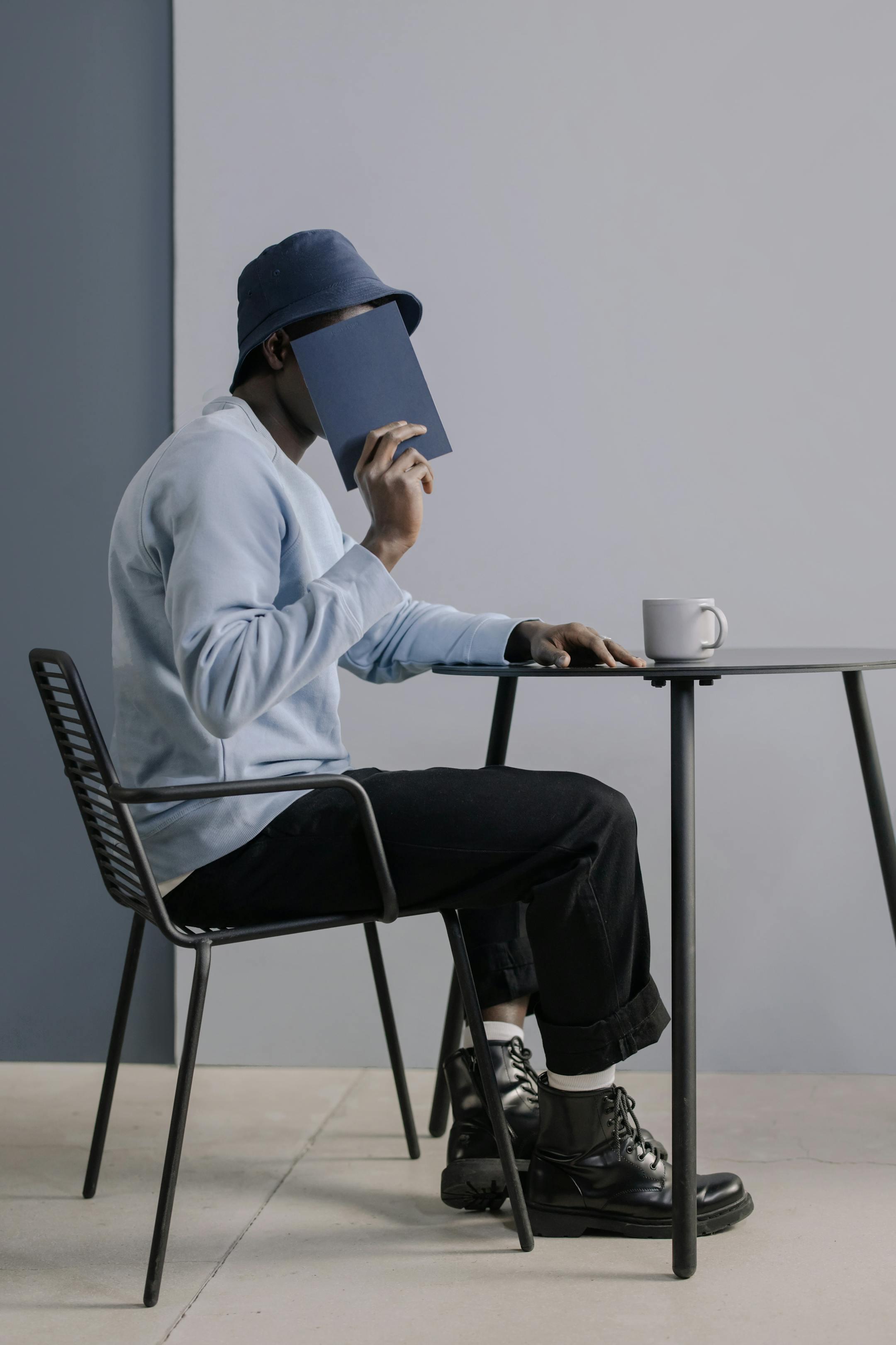 Fashionable man in a bucket hat sitting at a table, covering face with a book.