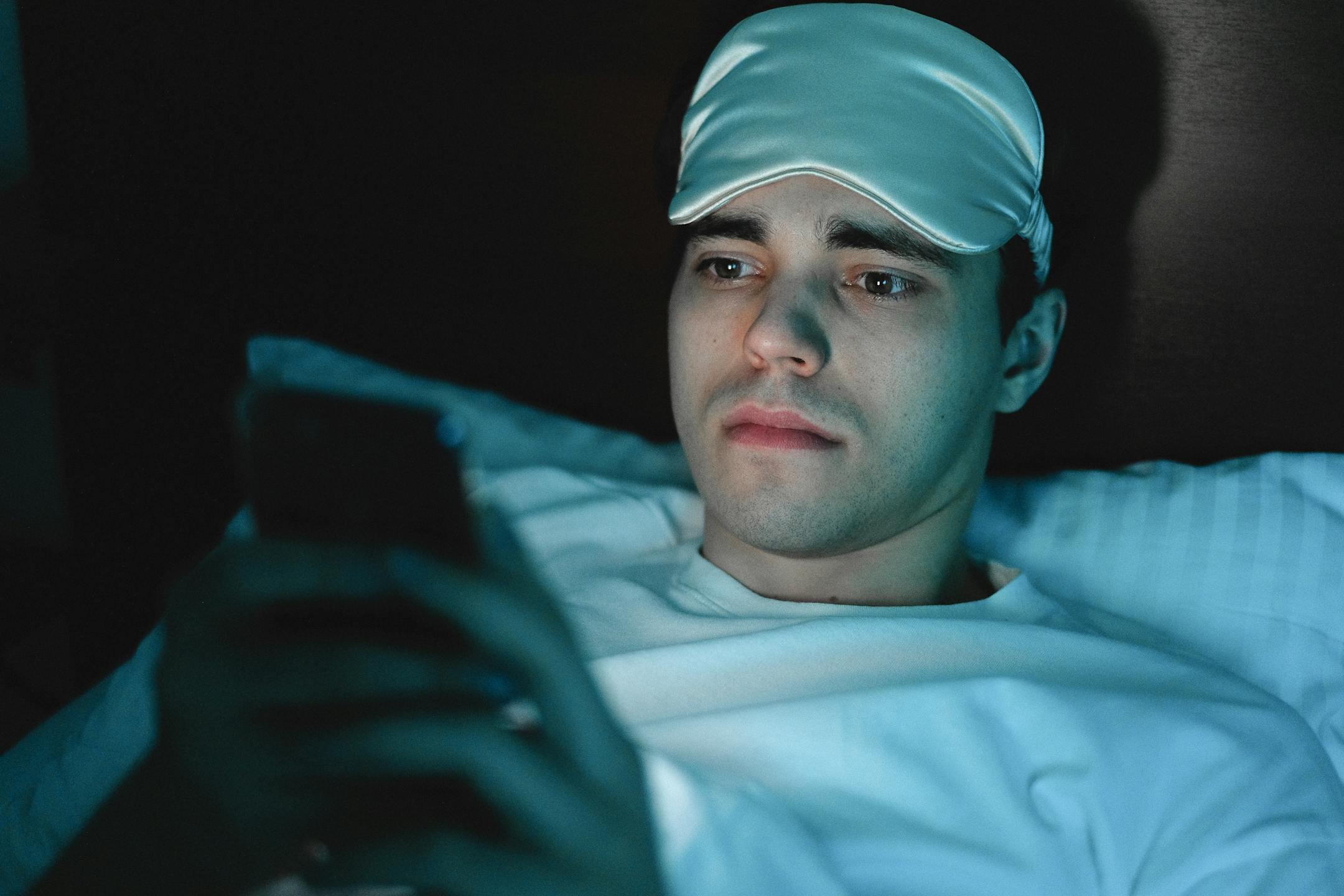 A man wearing a sleeping mask uses his smartphone while lying in bed at night.