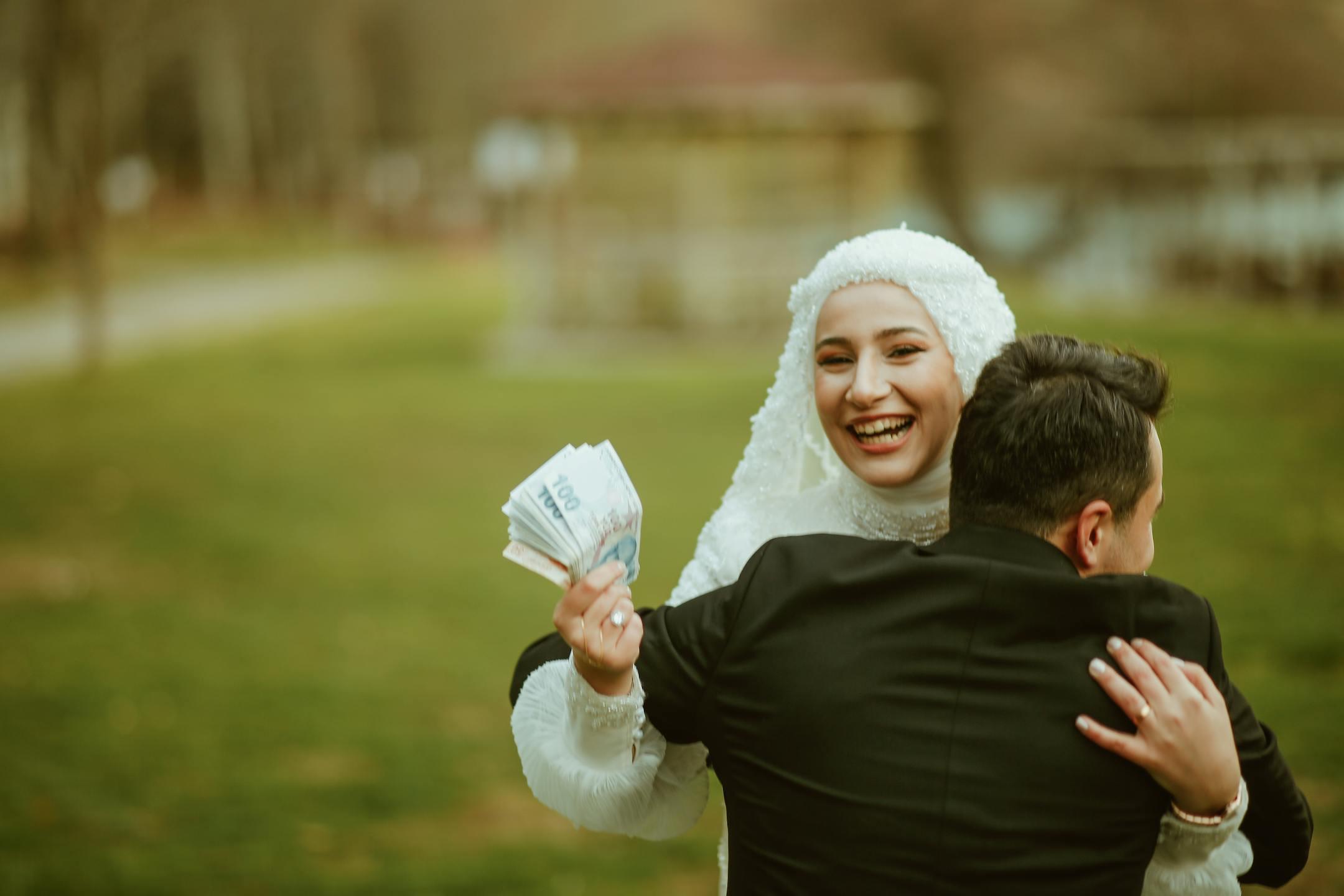 Happy bride in hijab holding money while embracing her groom outdoors.