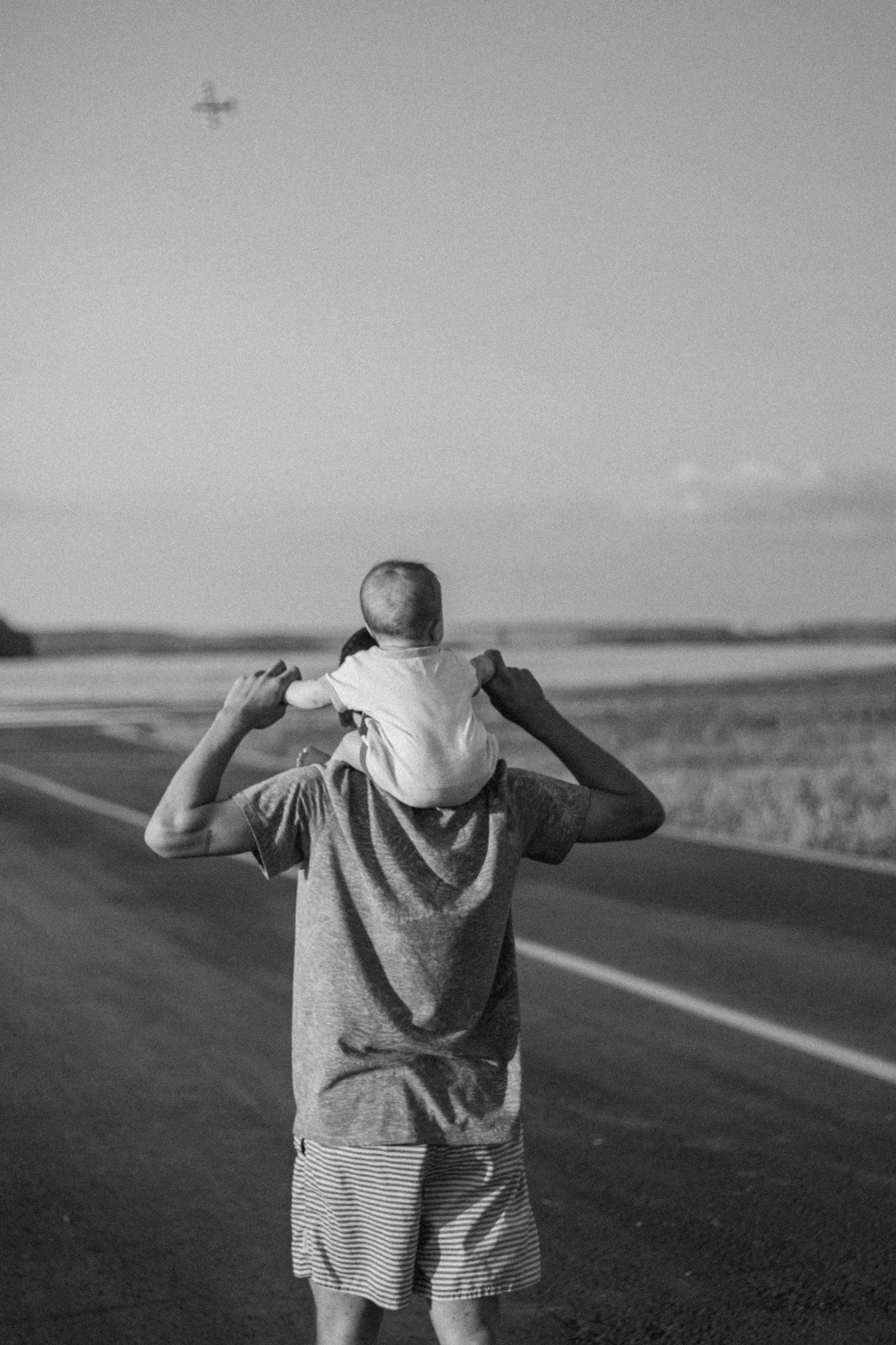 Black and white photo of a father carrying his child on his shoulders on a rural road.