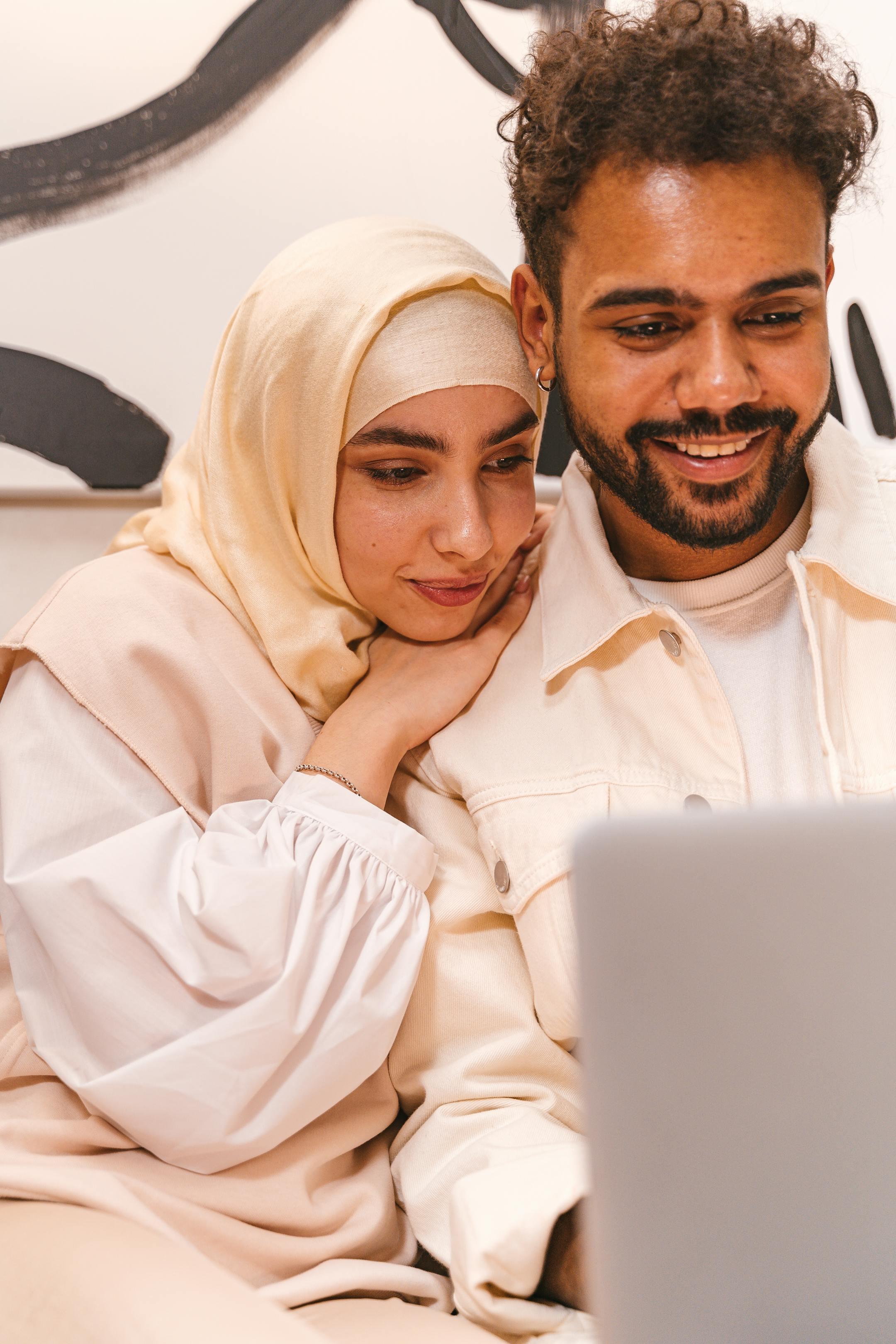 Muslim couple wearing hijab, sharing a warm moment while using a laptop indoors.
