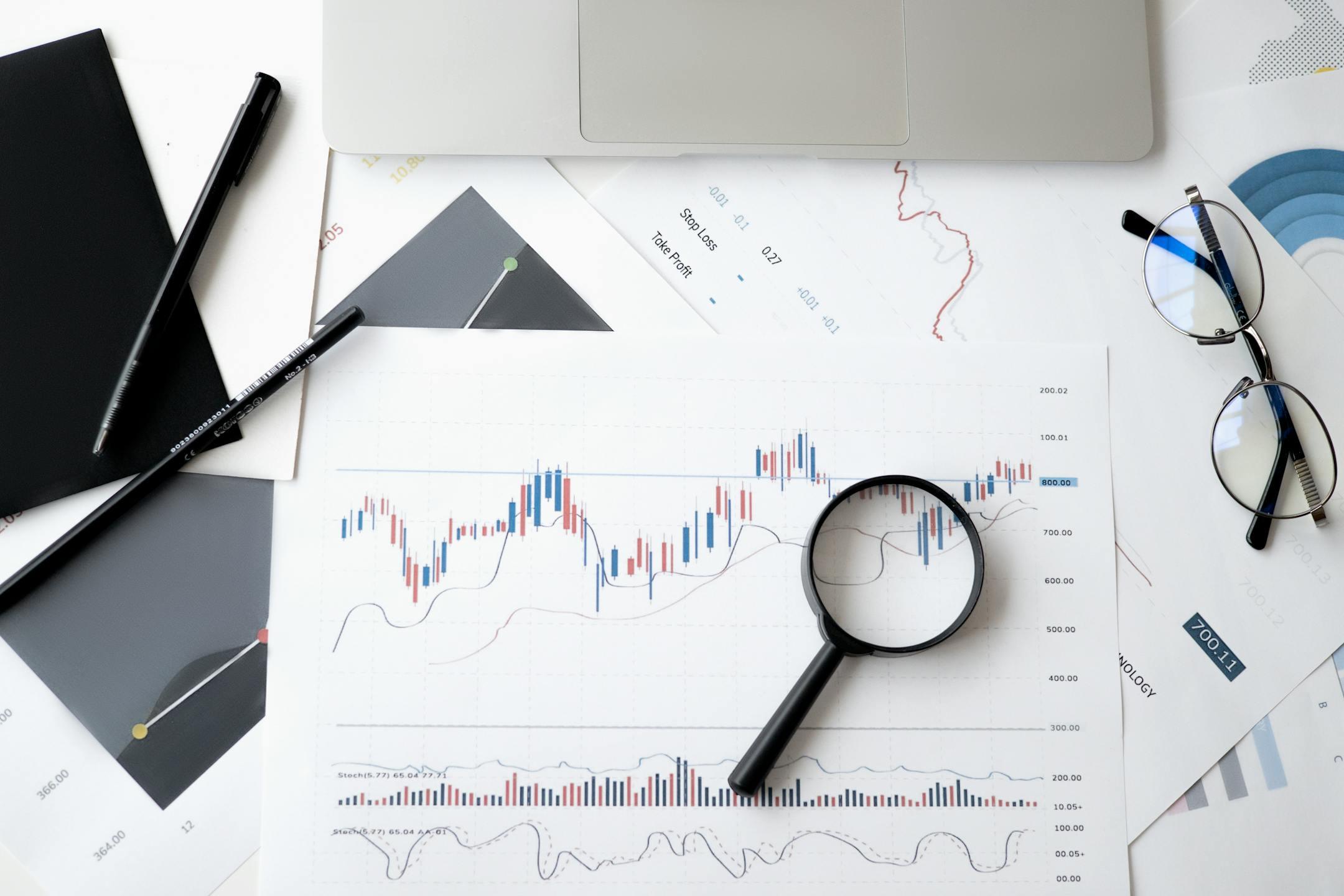 Flat lay of stock market analysis documents with a magnifying glass and glasses, symbolizing Cognitive Analytica’s focus on diagnosing and solving costly psychological phenomena that impact global economic performance.