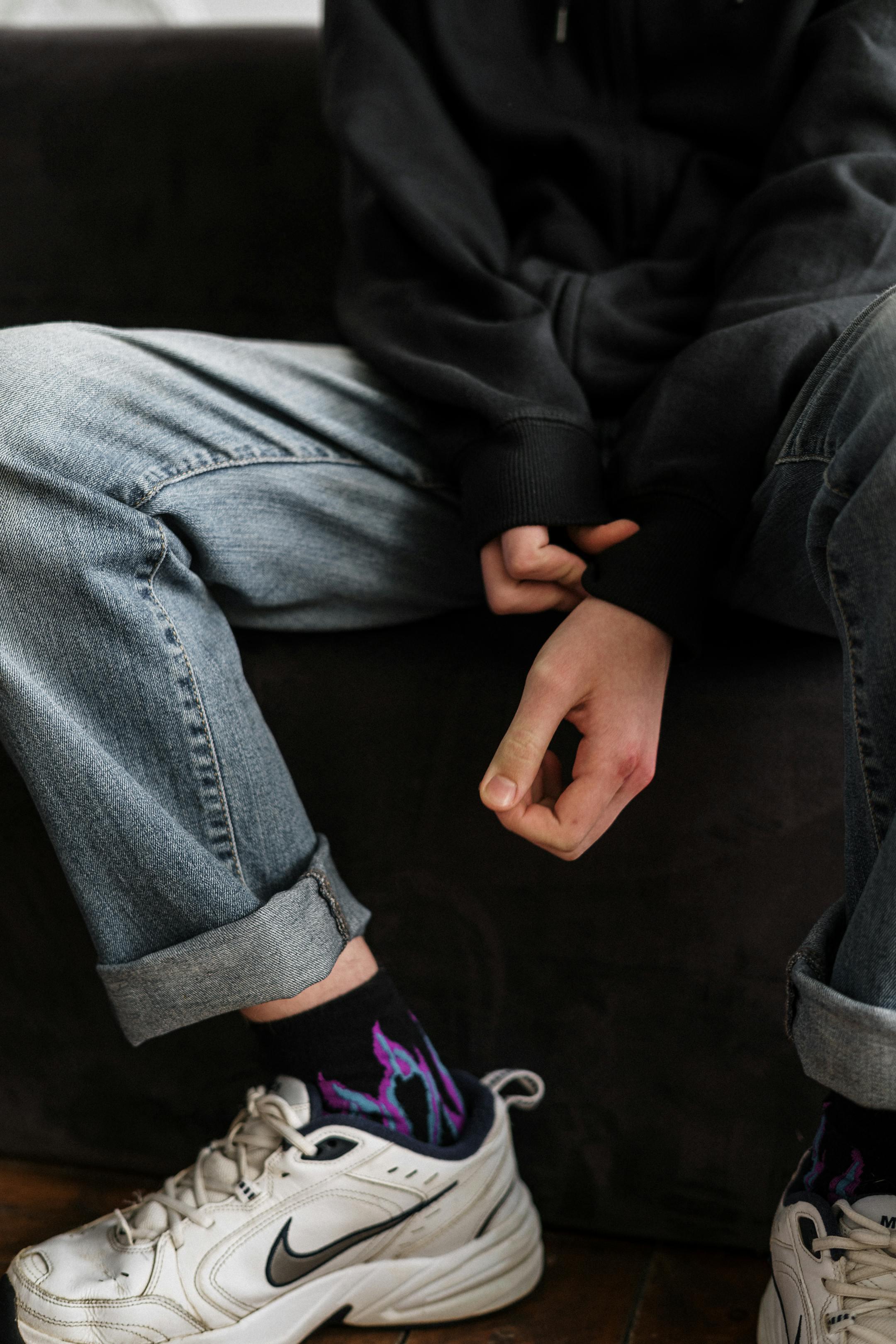 Teenager in a hoodie and jeans sitting casually indoors with crossed arms and white sneakers.