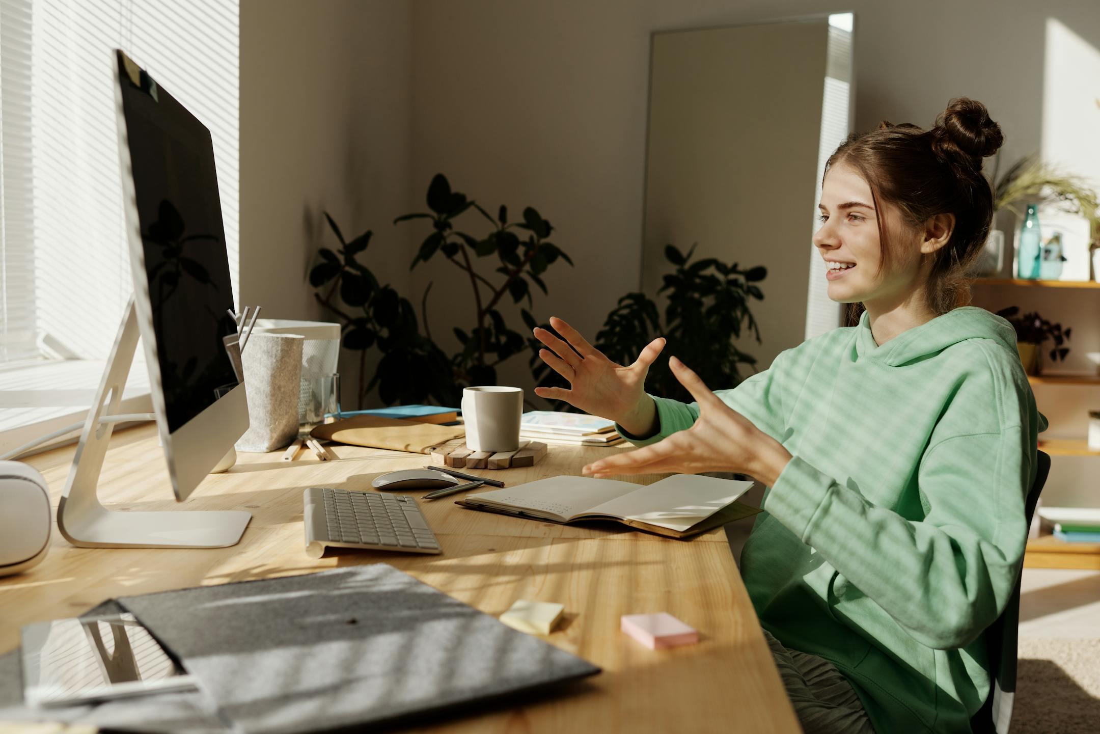 A woman in a green hoodie having a video call at her home office desk.