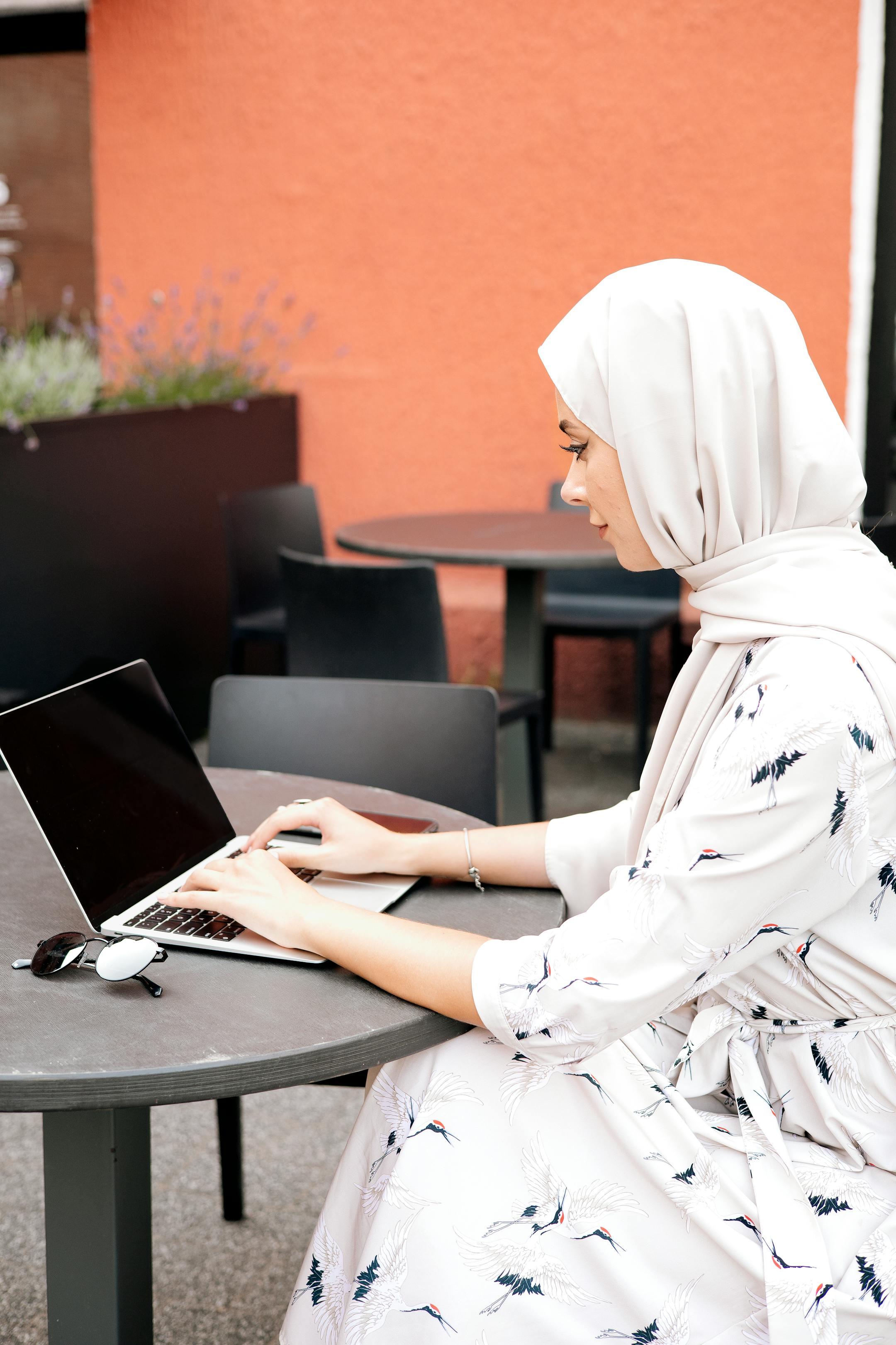 A woman in a hijab working on a laptop at an outdoor cafe table with a vibrant background.