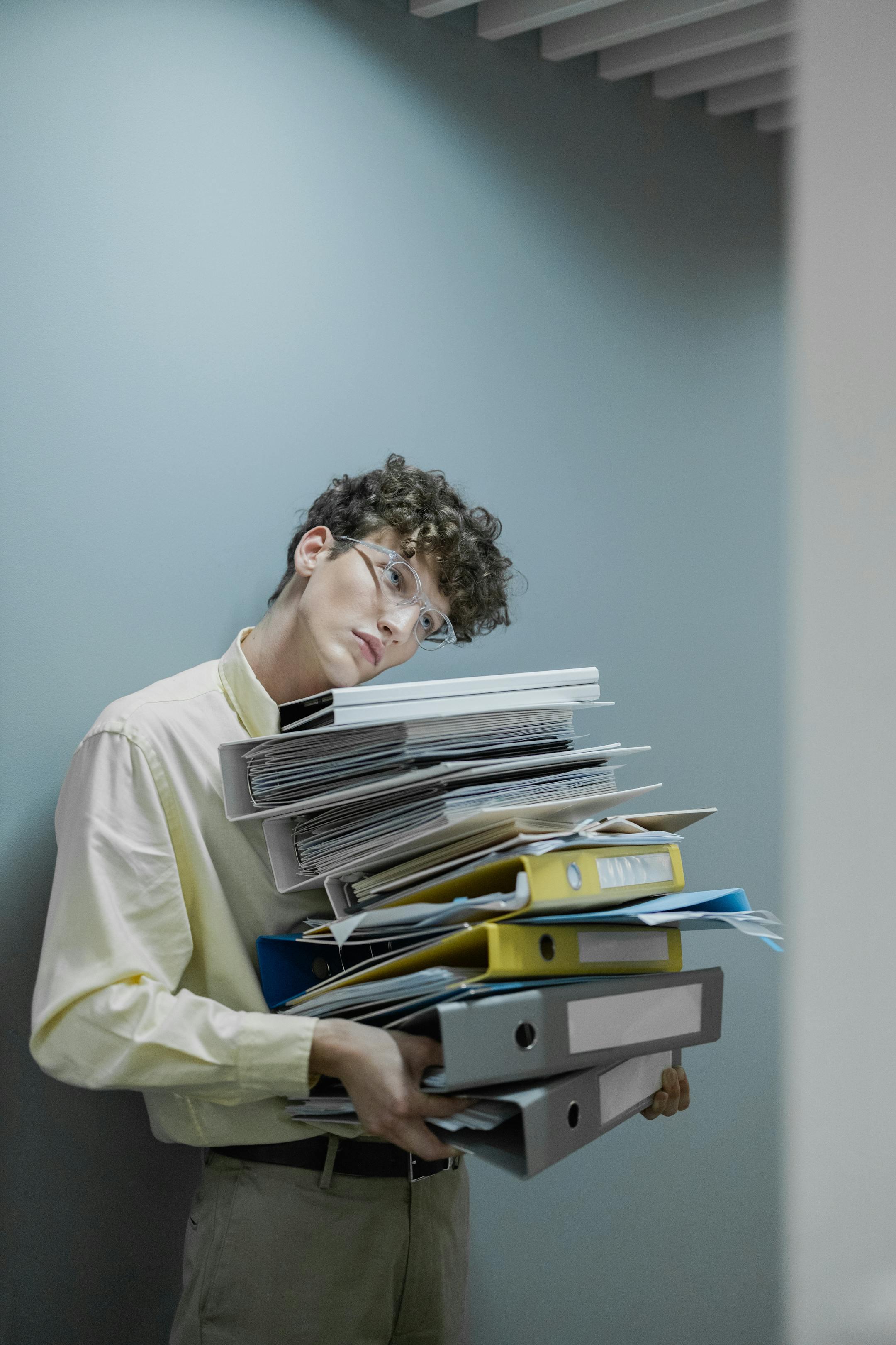 Young man with glasses carrying a stack of files looking tired. Office setting with gray wall.