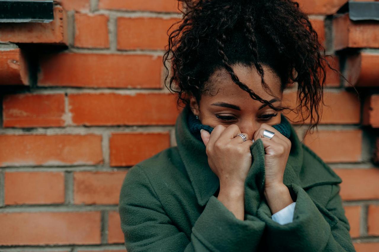 Woman in a green trench coat displays emotion, covering face with hands, against a brick wall.