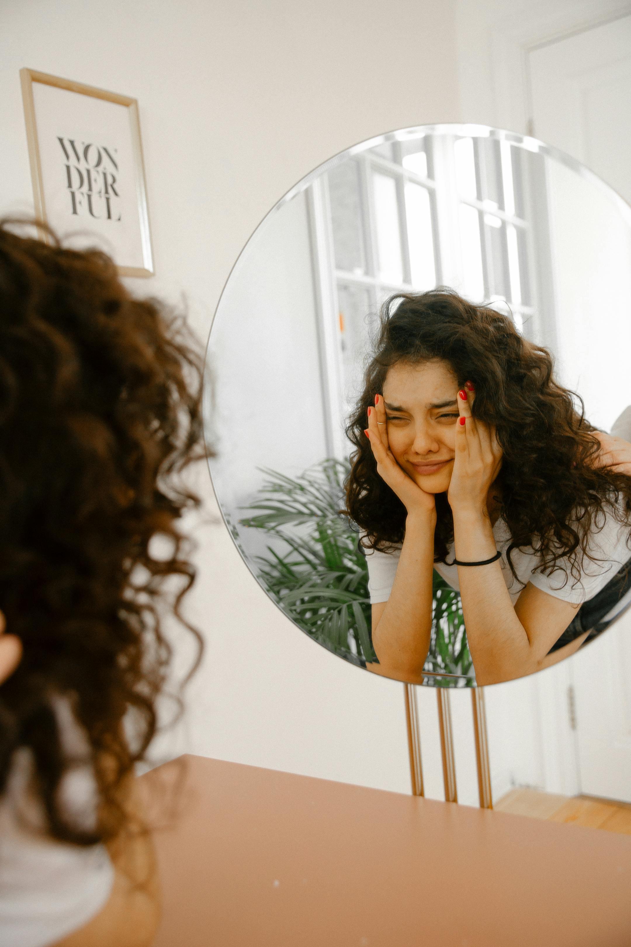Young woman with curly hair looking at her reflection in a round mirror, conveying mixed emotions.