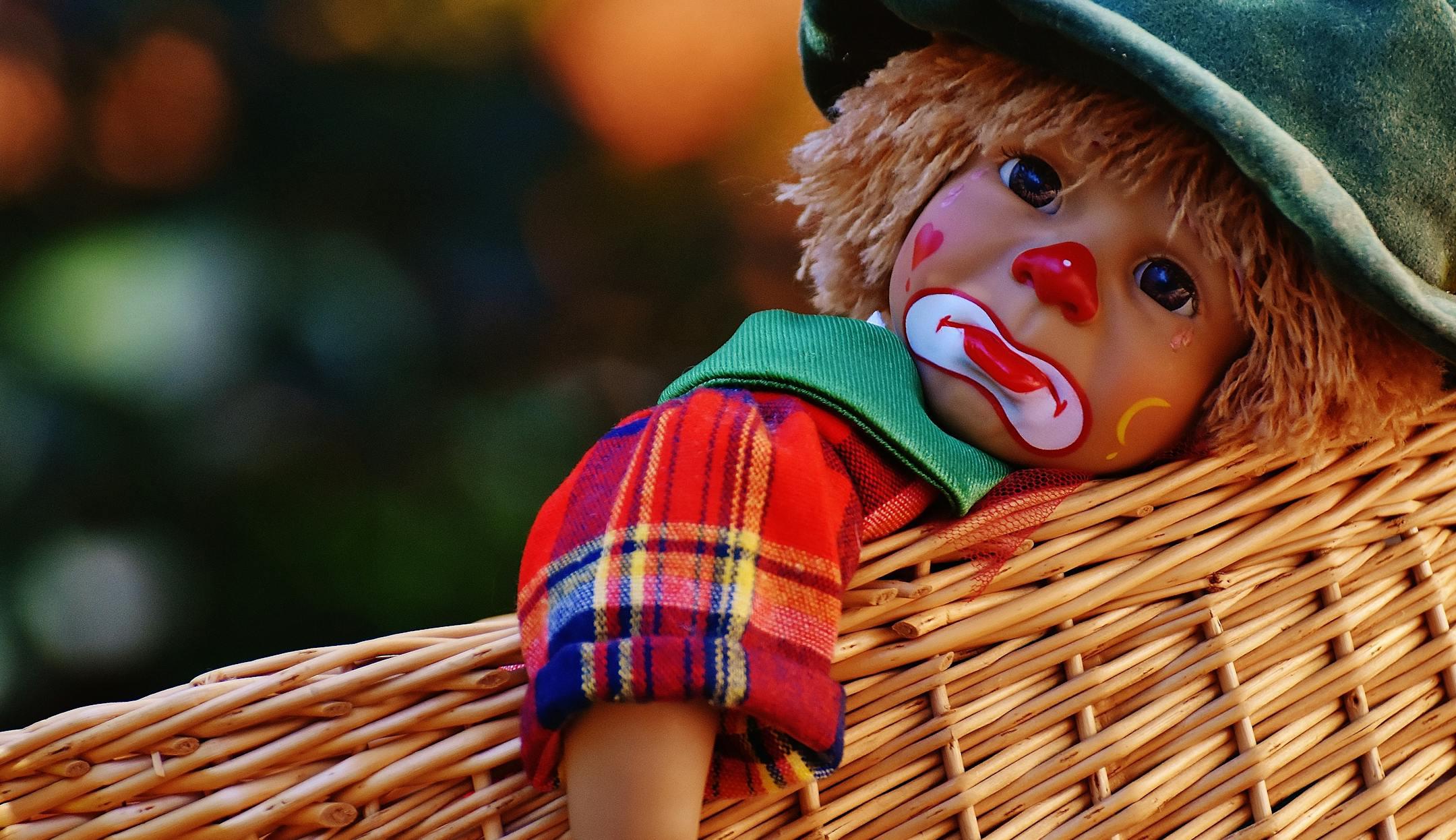 A close-up of a colorful clown doll with a sad expression lying in a woven basket.