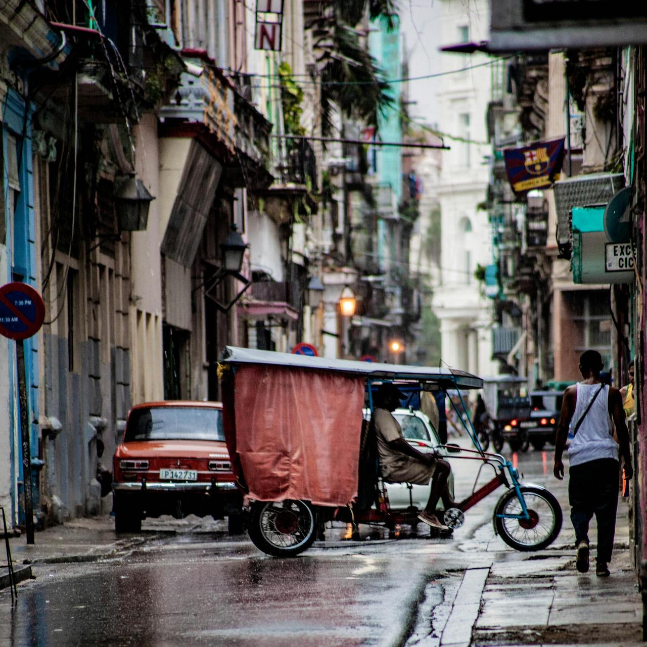 A bustling street in Havana, Cuba features classic cars, a rickshaw, and vibrant architecture.