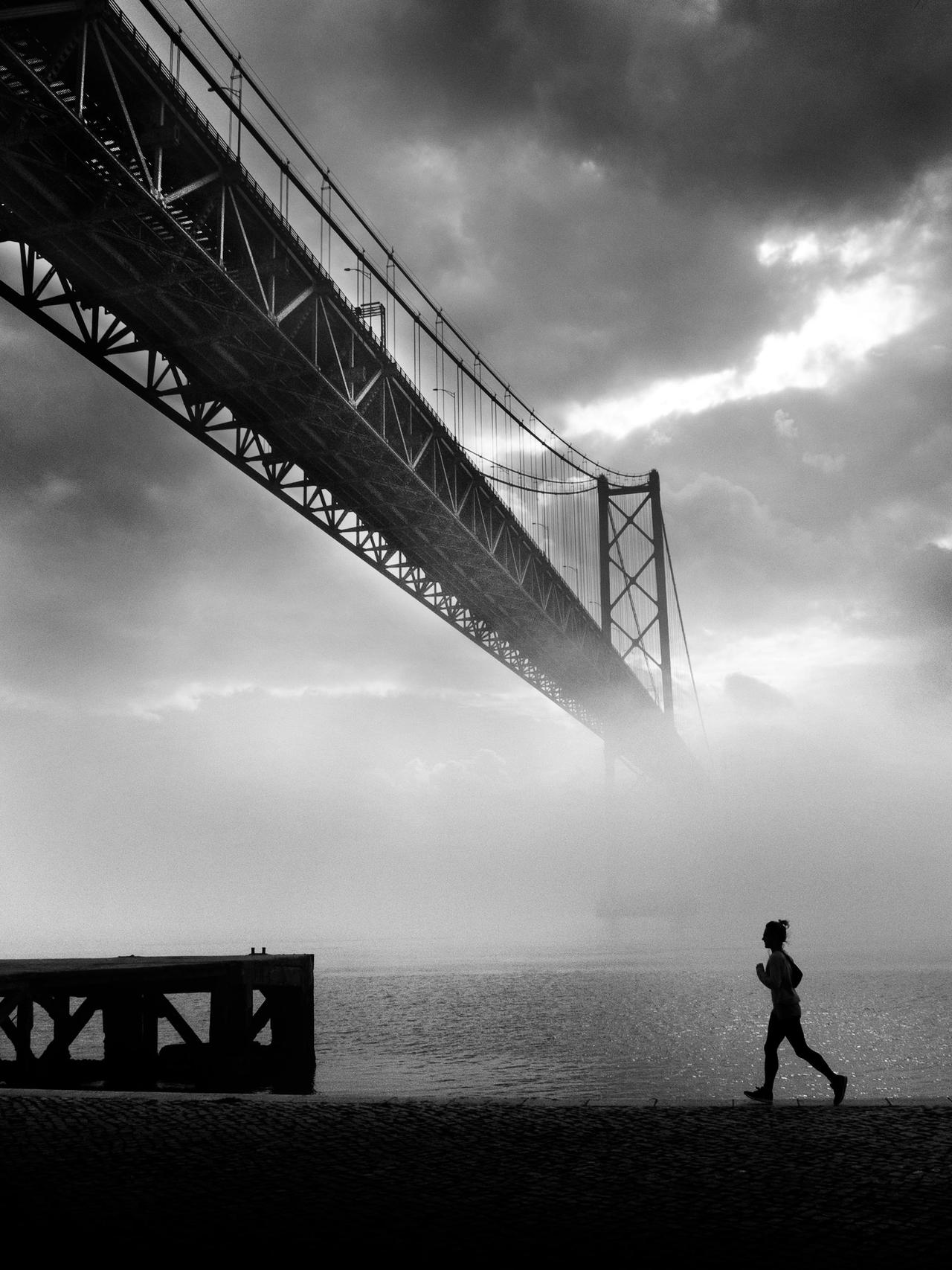 Silhouette of a person jogging near a misty river with a suspension bridge in the background.