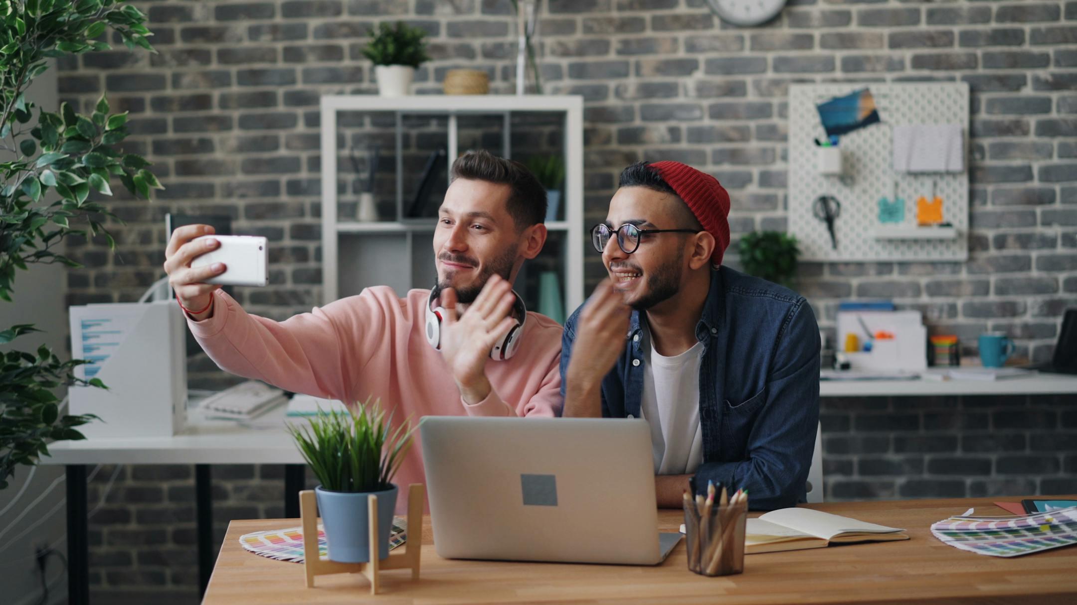 Two young men smiling and taking a selfie in a modern, creative office space.
