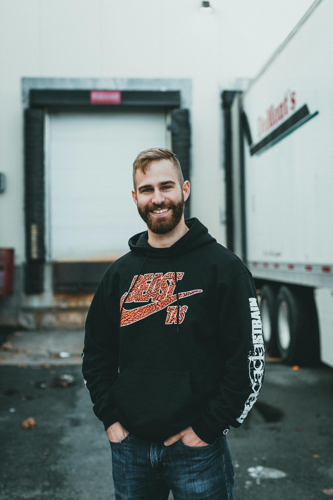 A smiling bearded man wearing a hoodie stands confidently outdoors by a truck.