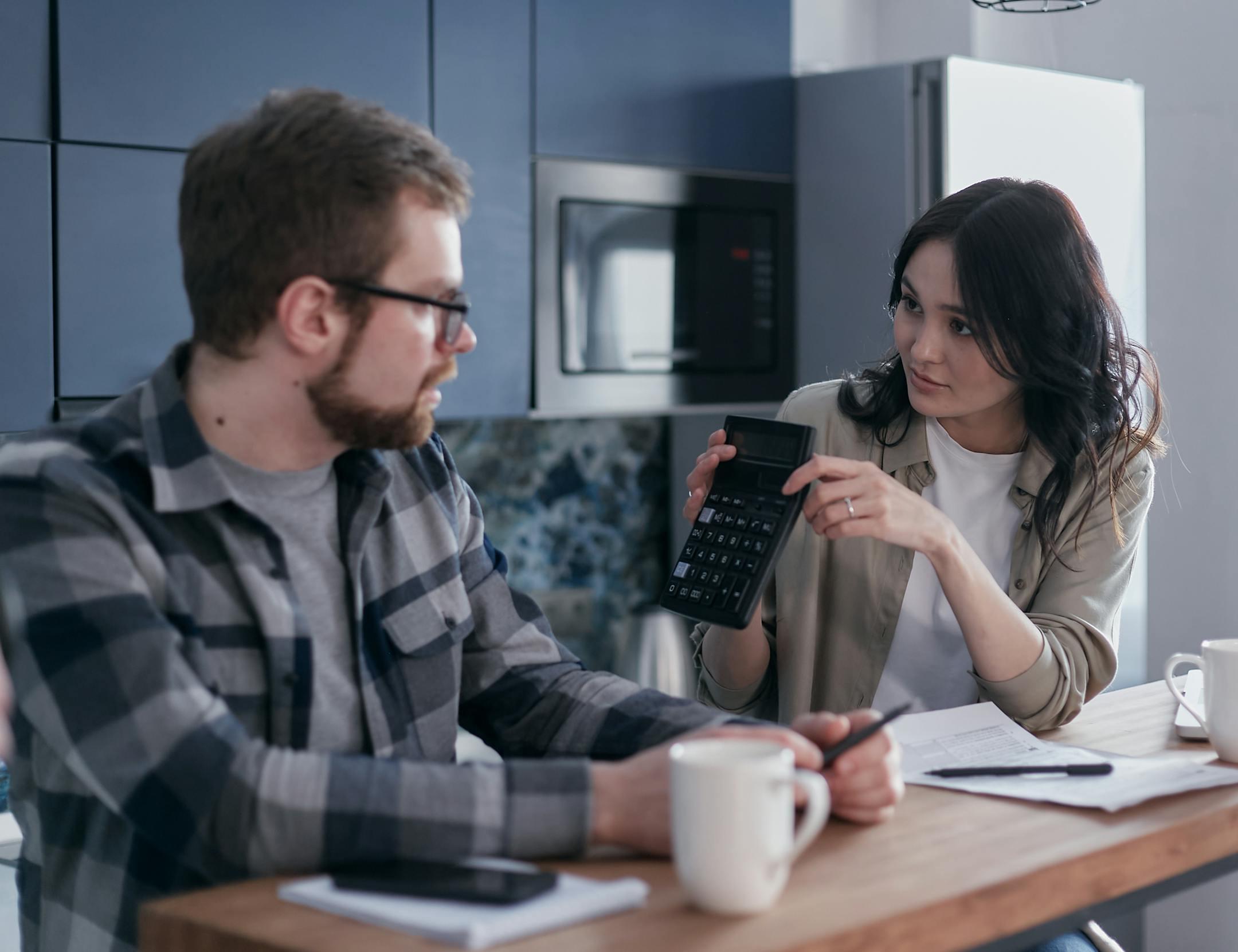 A couple discussing finances with a calculator and papers at a wooden table, looking concerned.