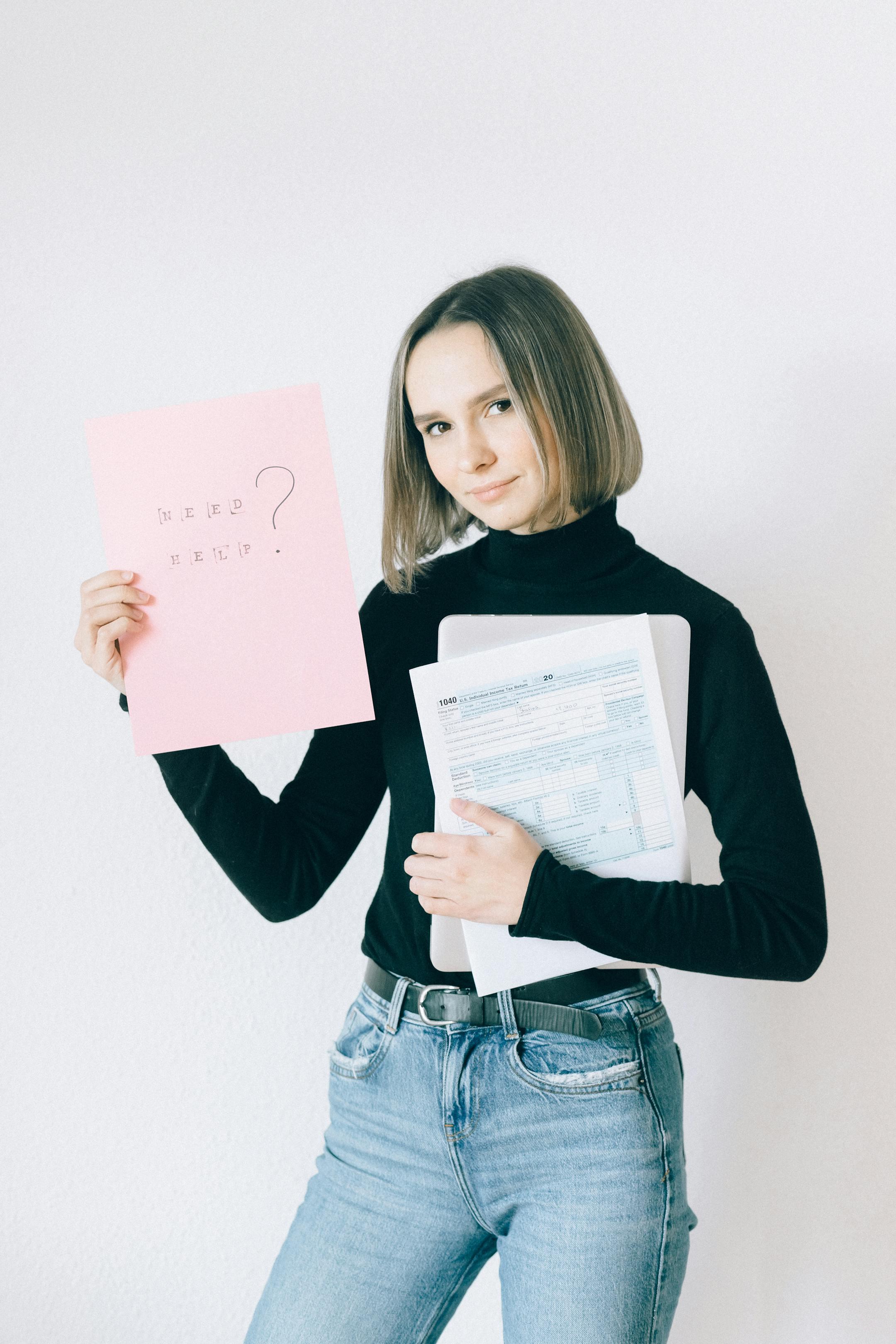 A woman in a black turtleneck holding tax forms and a pink paper, standing against a white background.