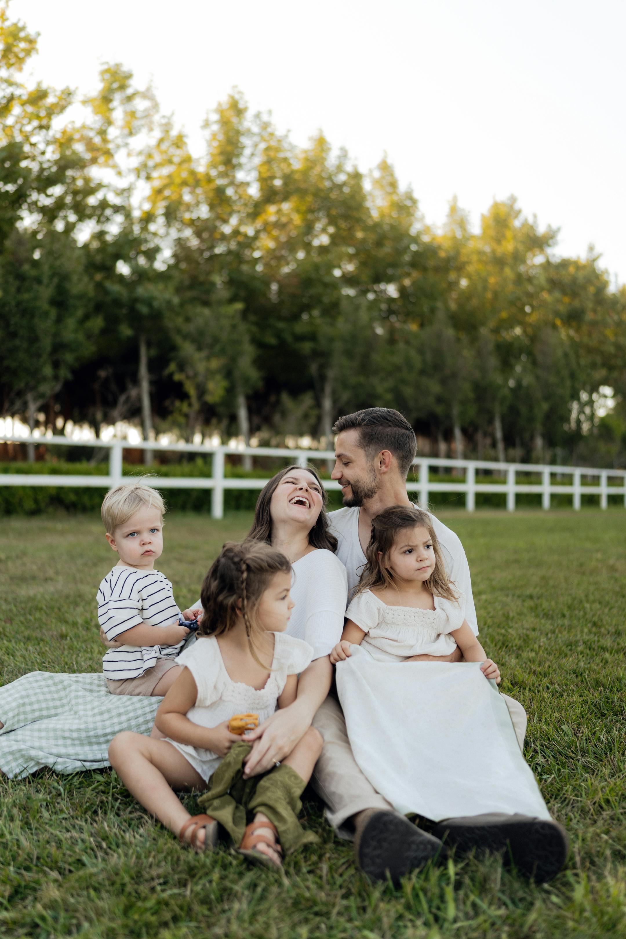 Family of five laughing and enjoying a sunny day on the grass with a forest background.