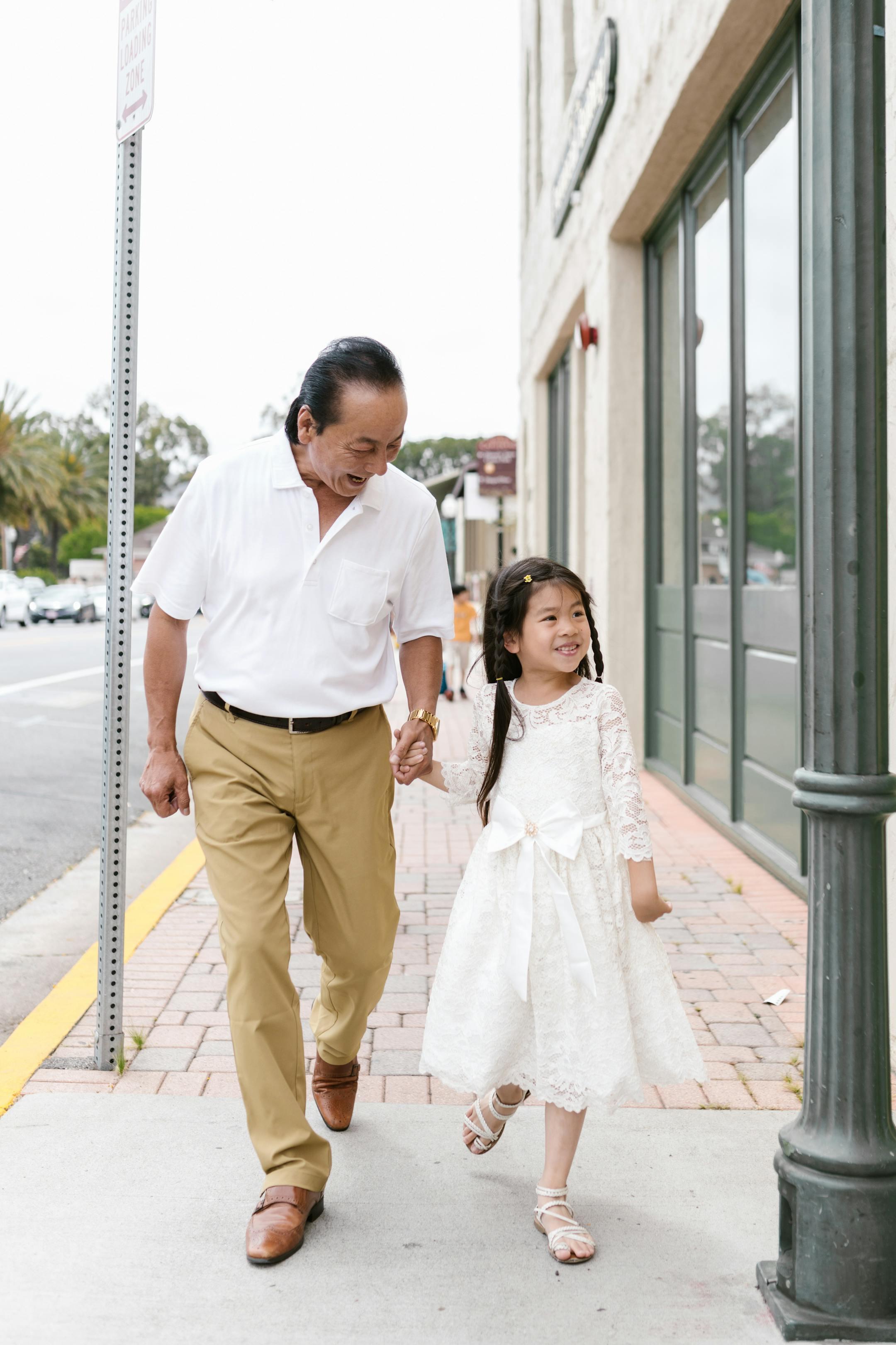 Elderly man and young girl holding hands and smiling while walking outdoors.