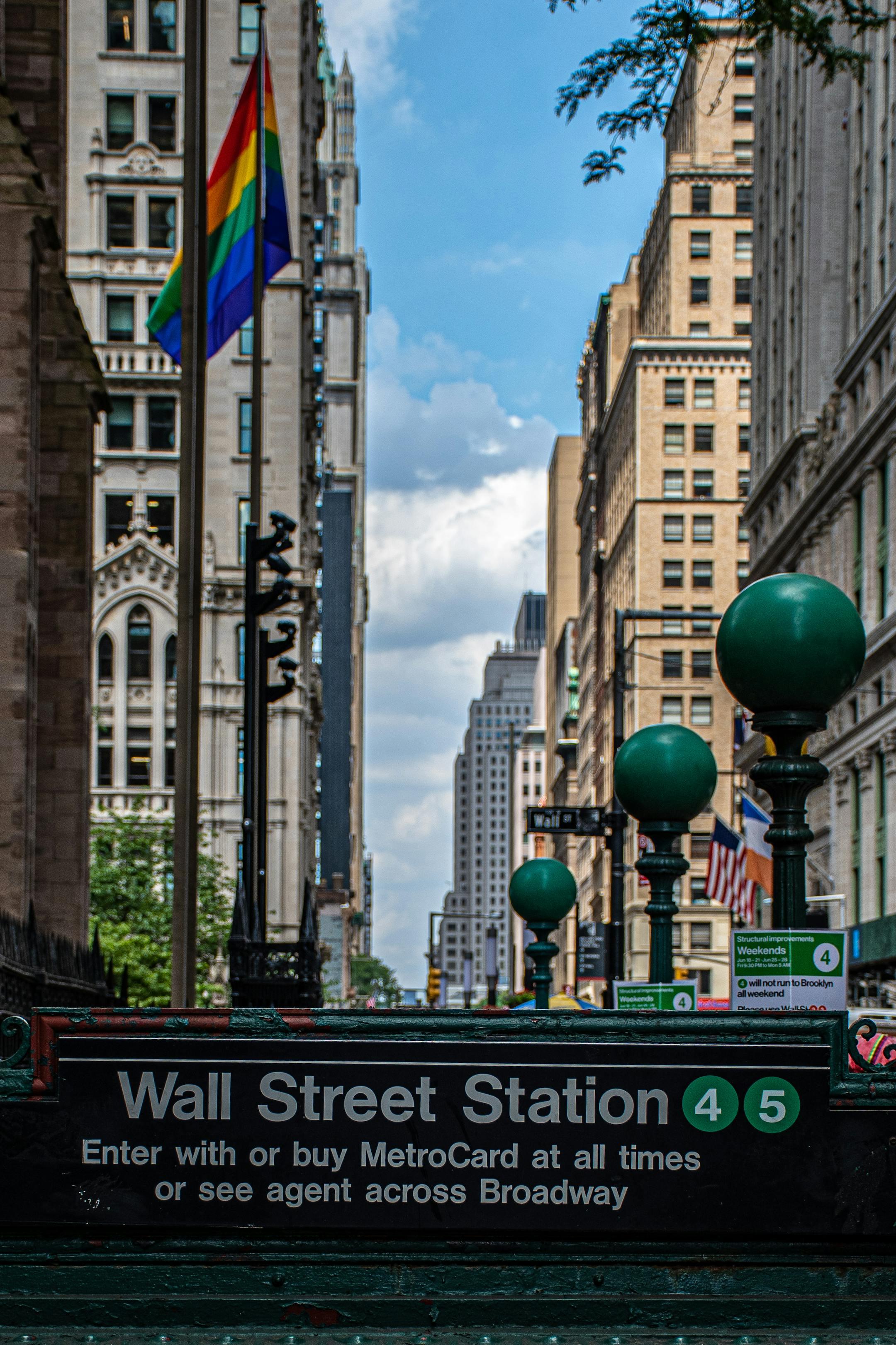 Urban scene of Wall Street Station entrance with skyscrapers and rainbow flag in NYC.
