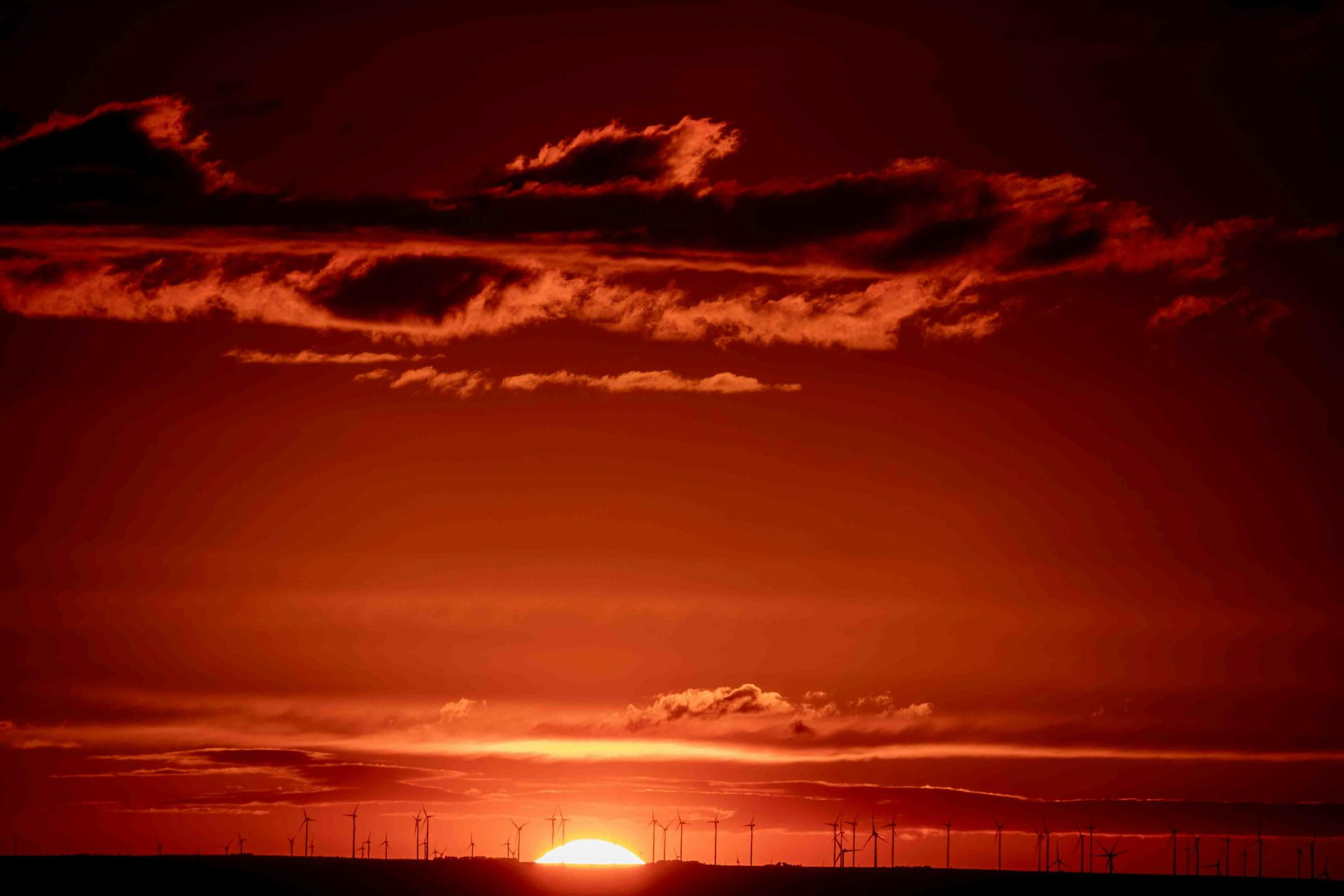 A dramatic sunset with the sun setting on the horizon, featuring the silhouettes of wind turbines against a fiery red sky, illustrating a fusion of natural beauty and renewable energy technology.