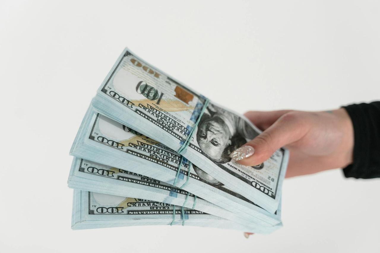 Close-up of a hand holding several stacks of US hundred dollar bills on a white background.