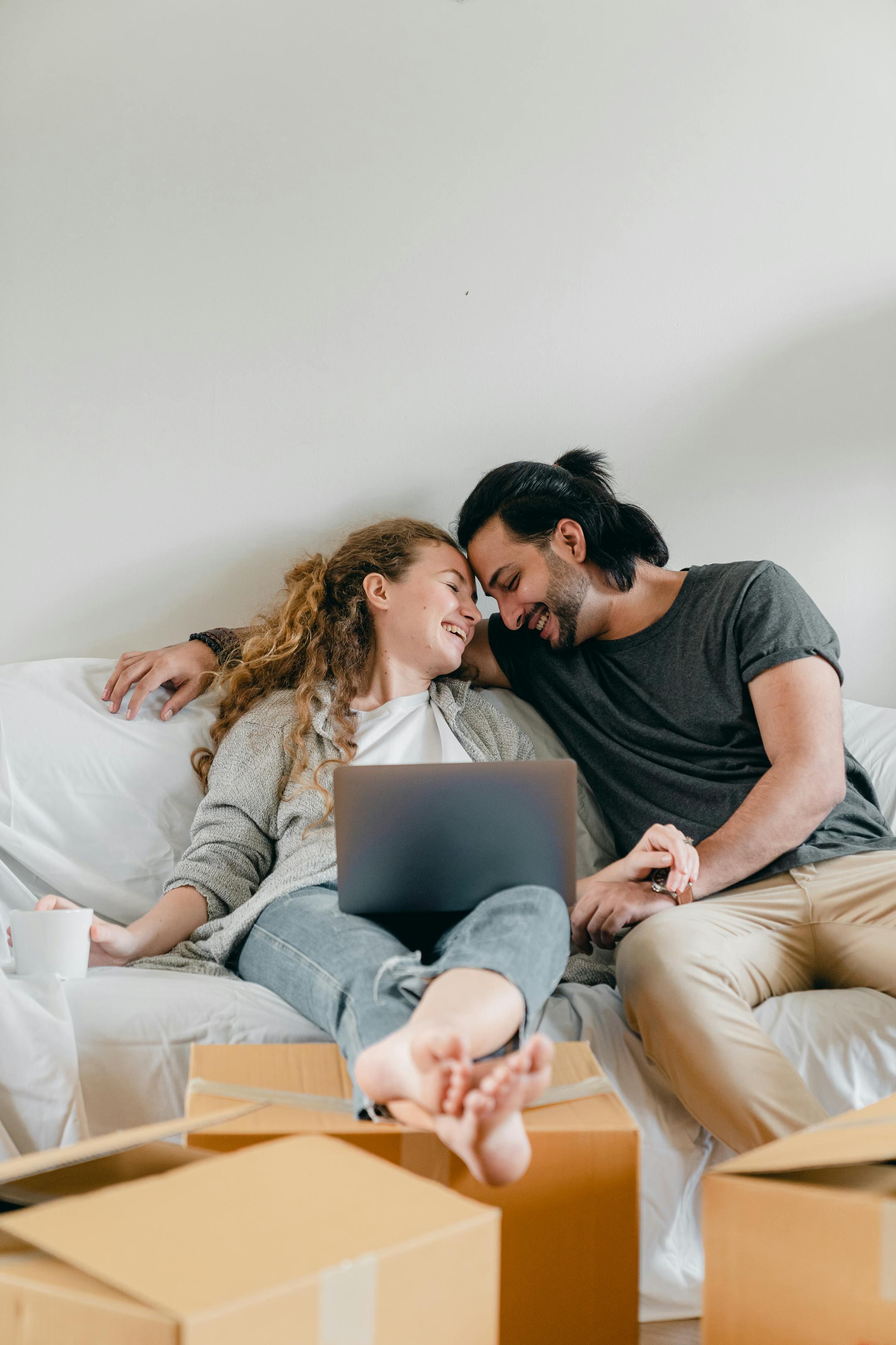 Positive barefoot woman in jeans with netbook sitting with crossed legs near happy ethnic boyfriend with closed eyes on cozy couch near cardboard boxes in living room