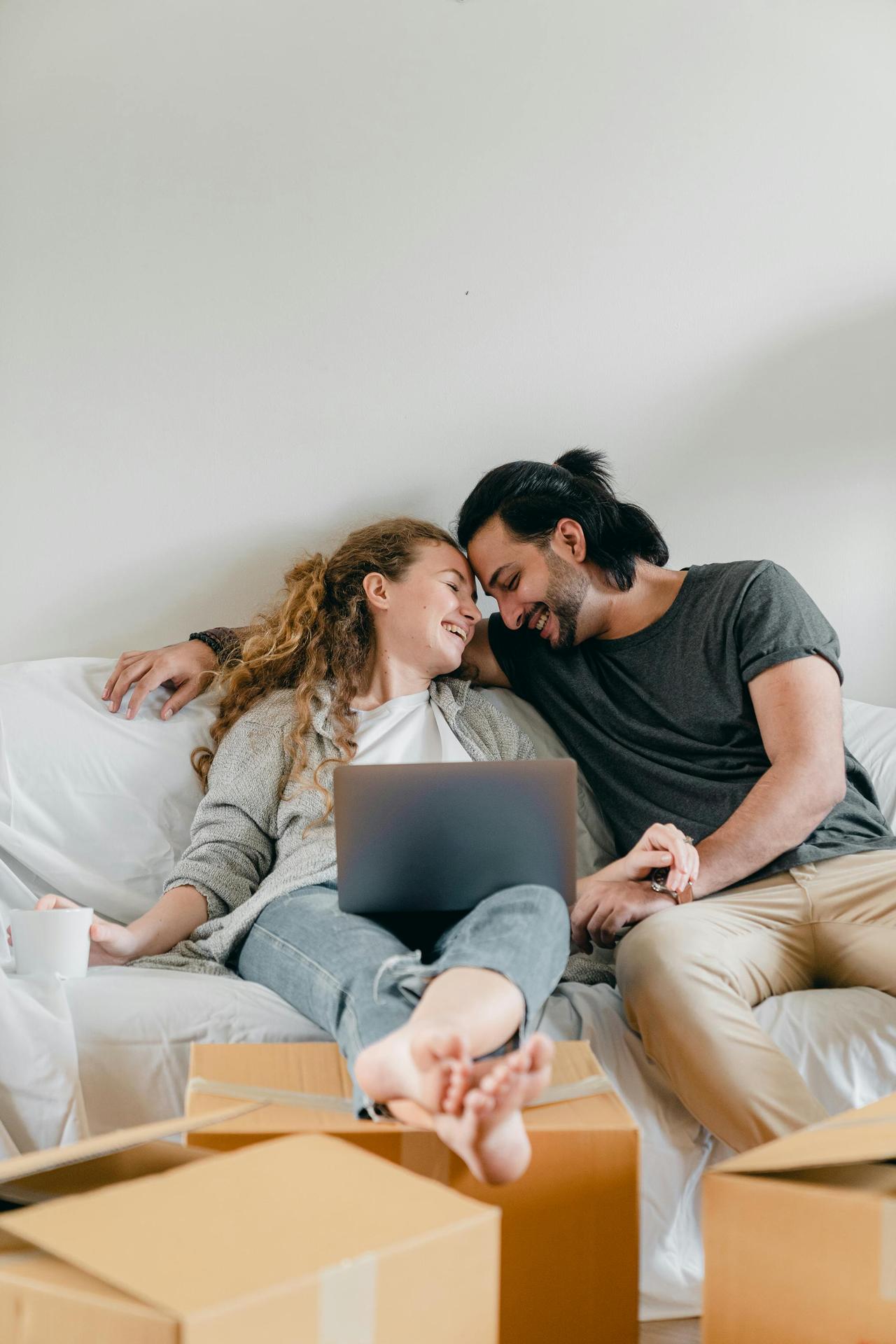 Positive barefoot woman in jeans with netbook sitting with crossed legs near happy ethnic boyfriend with closed eyes on cozy couch near cardboard boxes in living room