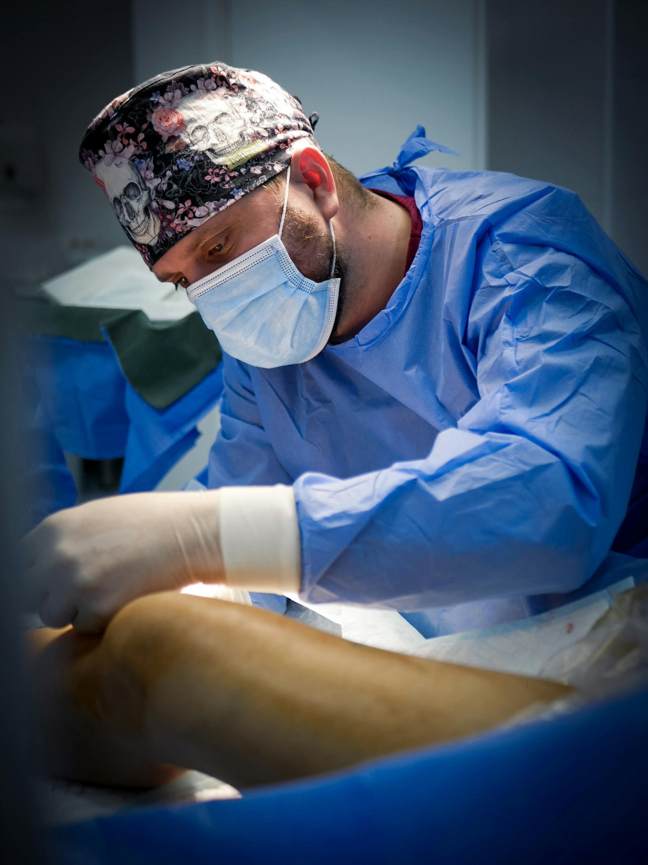 Surgeon in blue uniform and face mask performing a surgery in a hospital operating room.