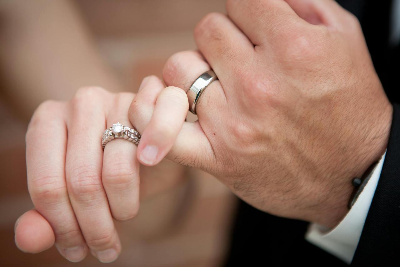 A close-up photo of a couple's hands interlocked, wearing wedding and engagement rings.