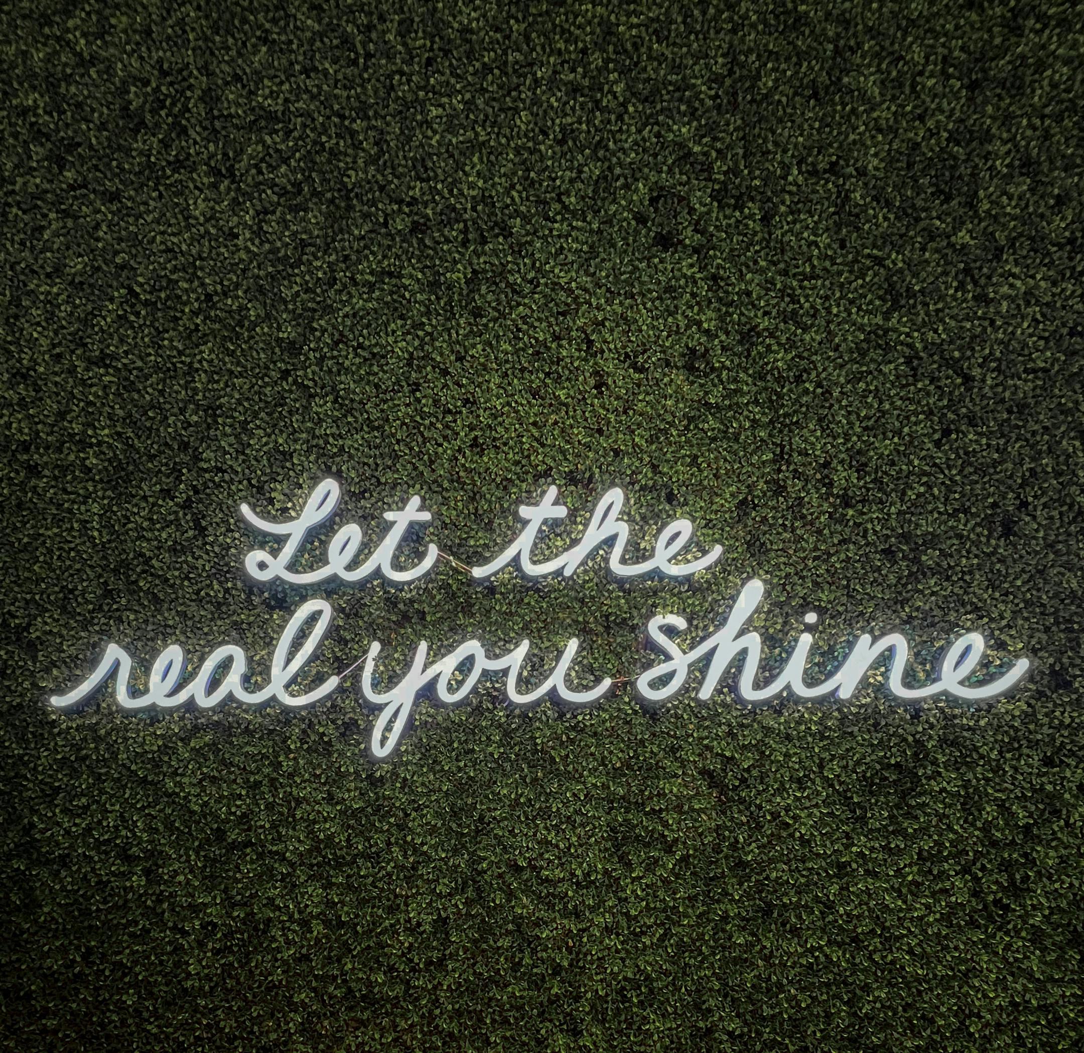Neon sign reading 'Let the real you shine' against a green textured wall, offering motivation.