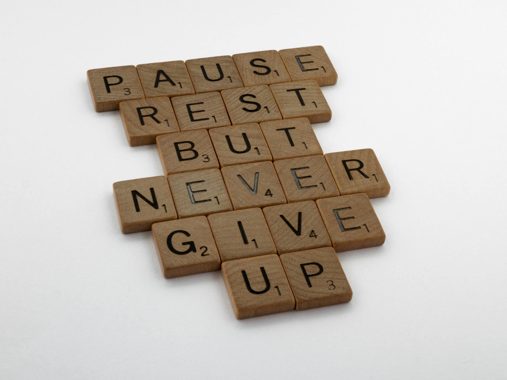 Wooden tiles spelling out 'Pause Rest But Never Give Up' on a white background.