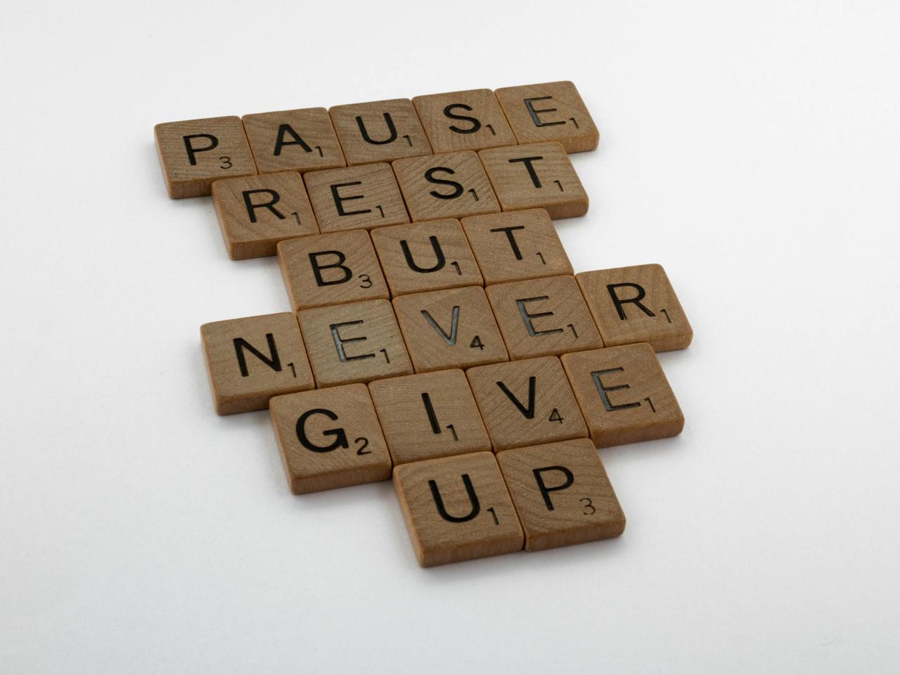 Wooden tiles spelling out 'Pause Rest But Never Give Up' on a white background.