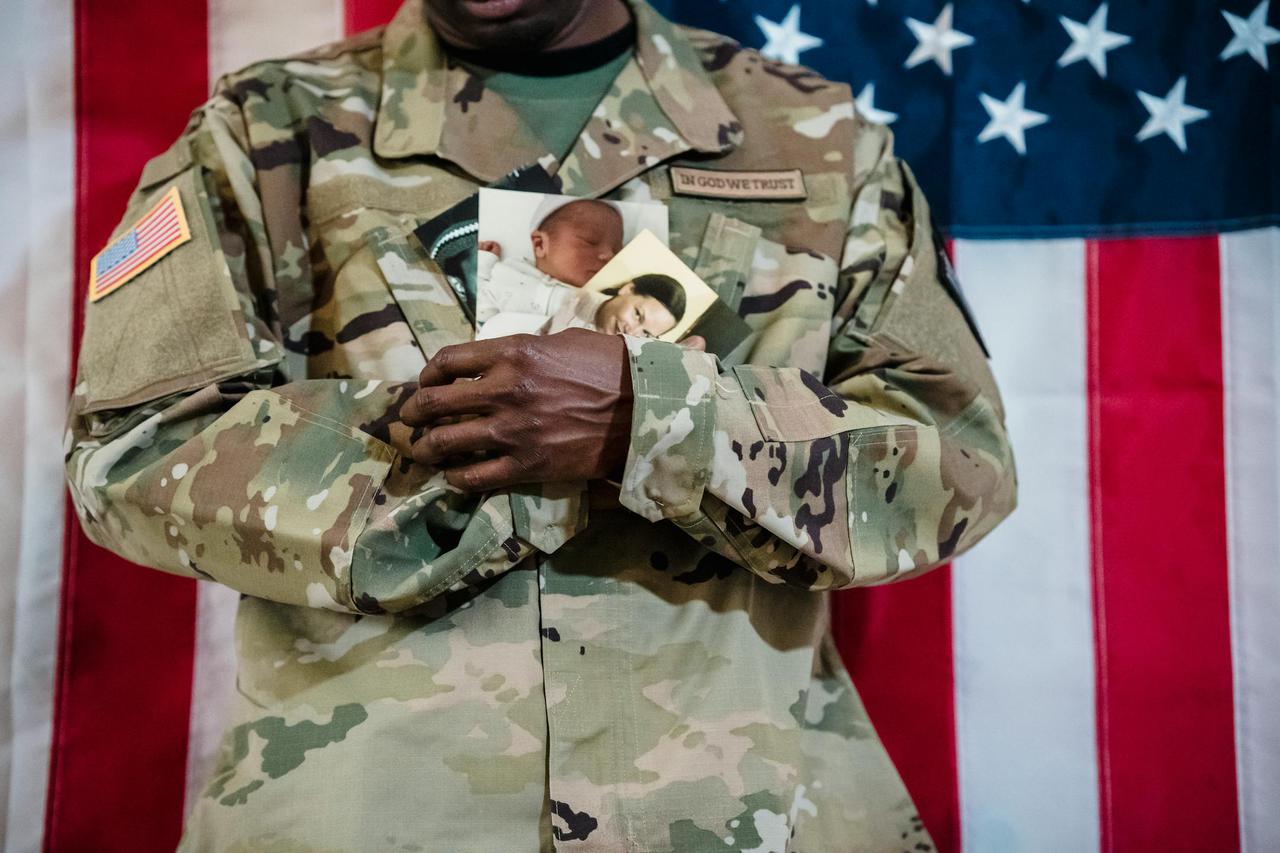 American soldier in uniform holding family photos against a flag backdrop.
