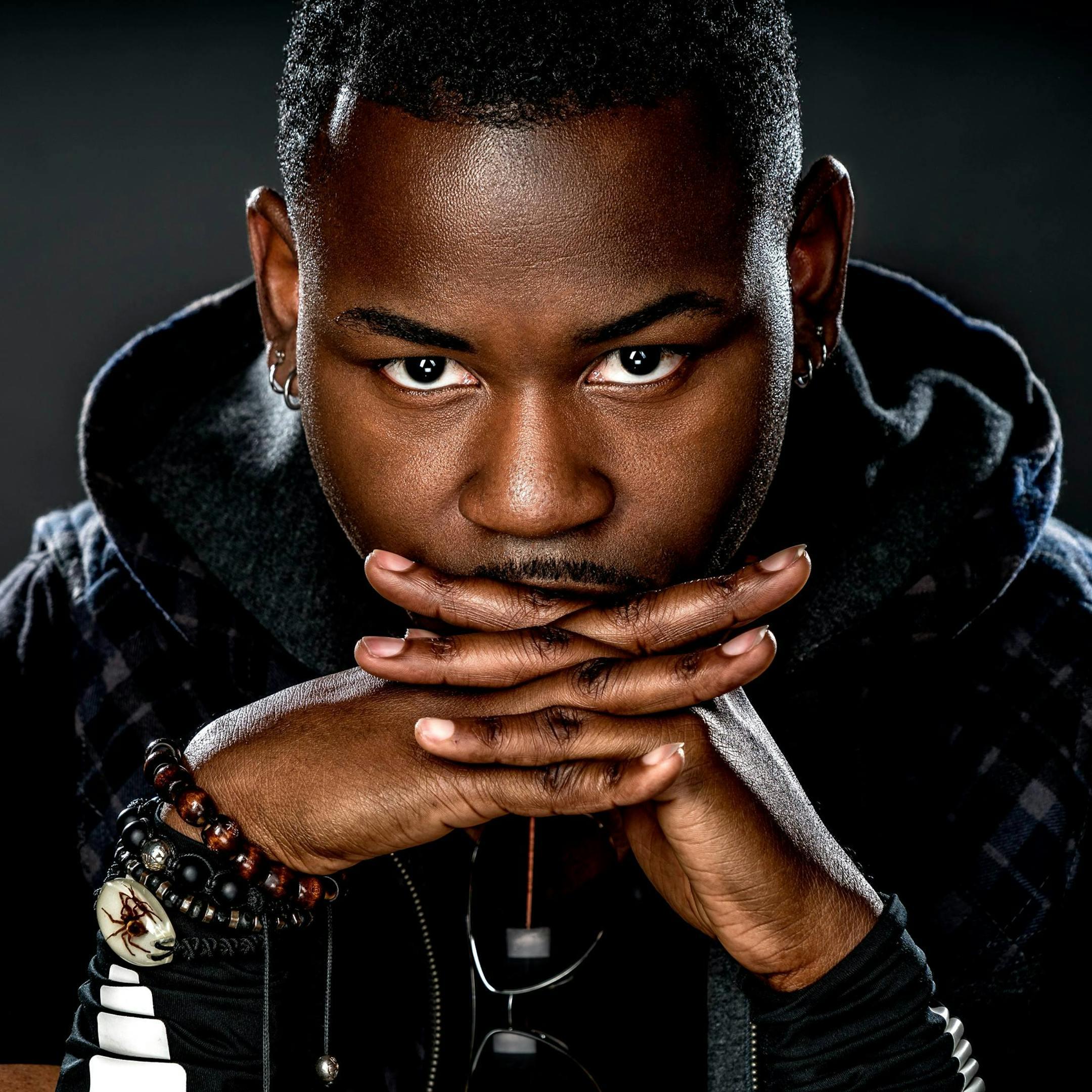 Close-up portrait of a young black man with hands clasped in a studio setting.
